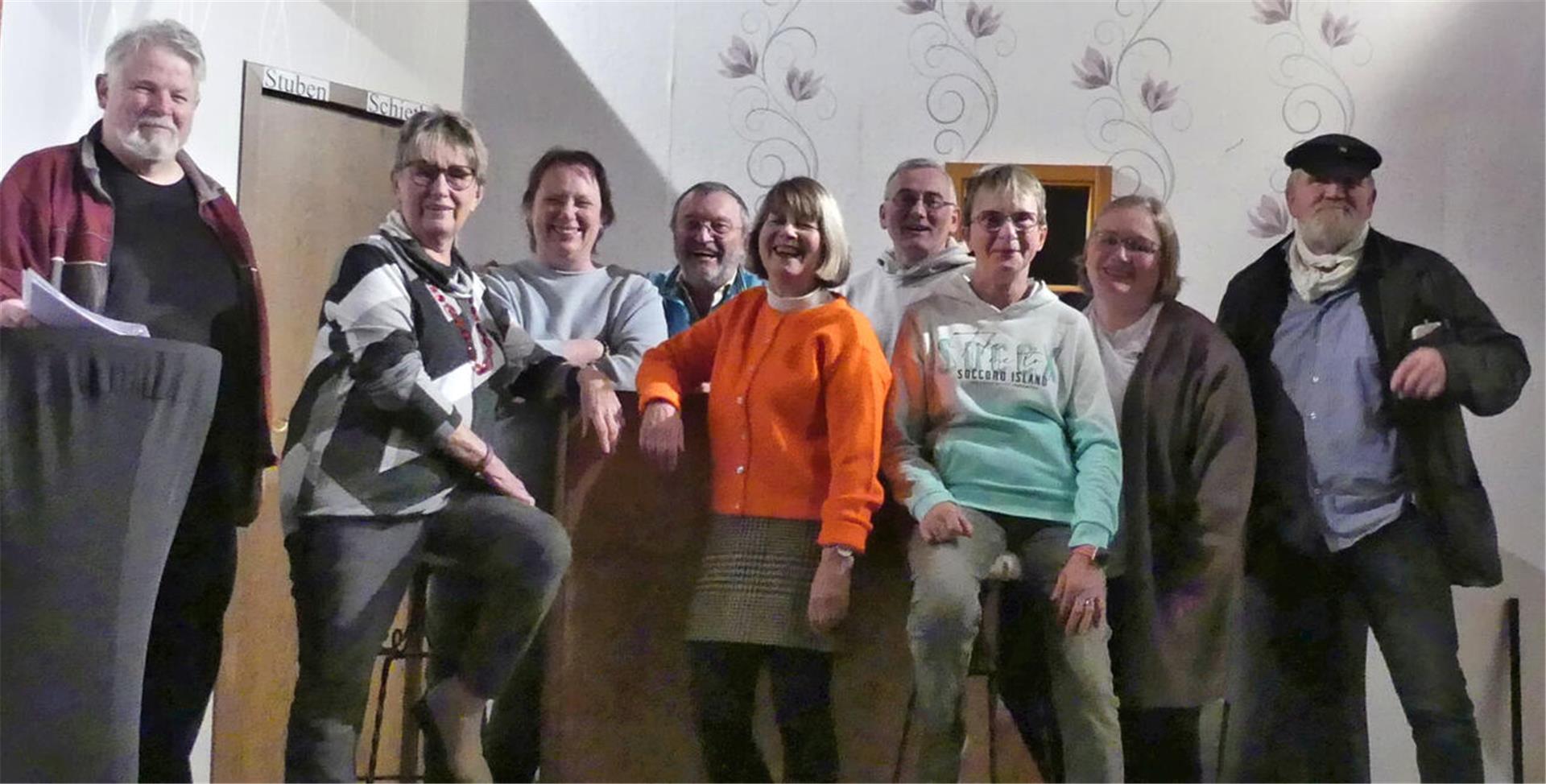 Ein Gruppenfoto von neun Personen, die in einer entspannten Pose in einem Innenraum zusammenstehen. Im Hintergrund ist eine helle Wand mit Blumenmustern und einer Holztür zu sehen. Die Personen wirken freundlich und lächelnd, einige lehnen an einer Theke oder einem Tisch.