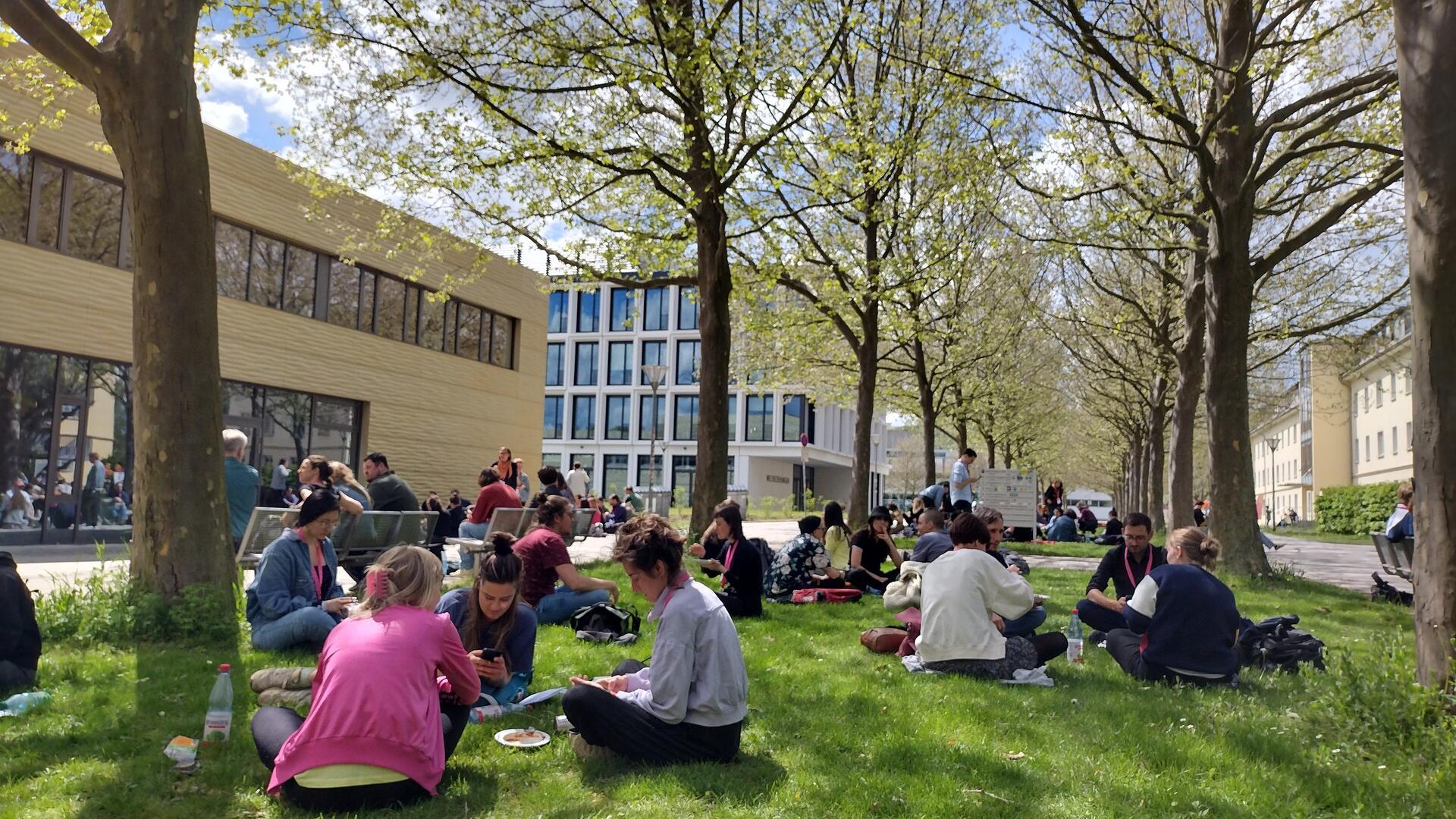 Viele junge Menschen sitzen auf einer Wiese zwischen Universitätsgebäuden.