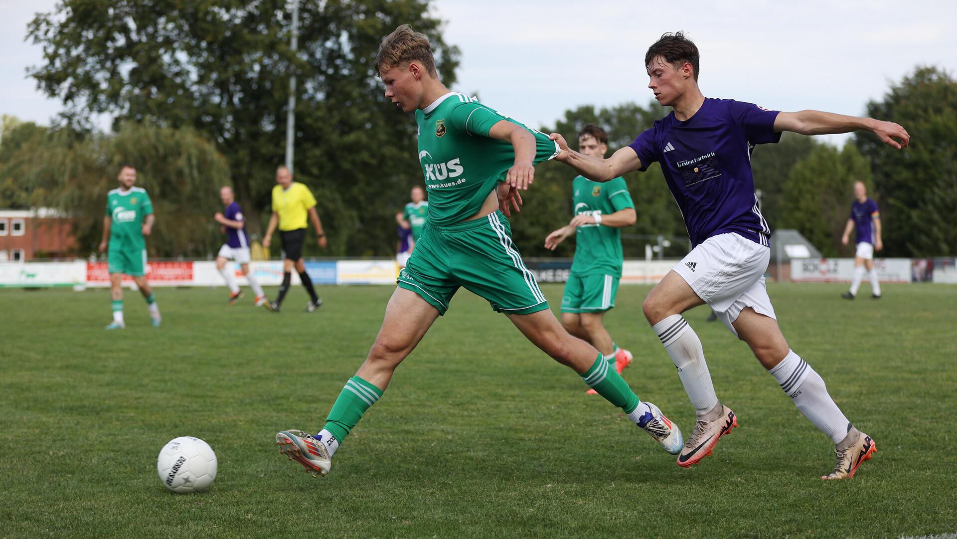 Zwei Männer laufen hinter einem Ball her, der hintere zieht den vorderen am Trikot.