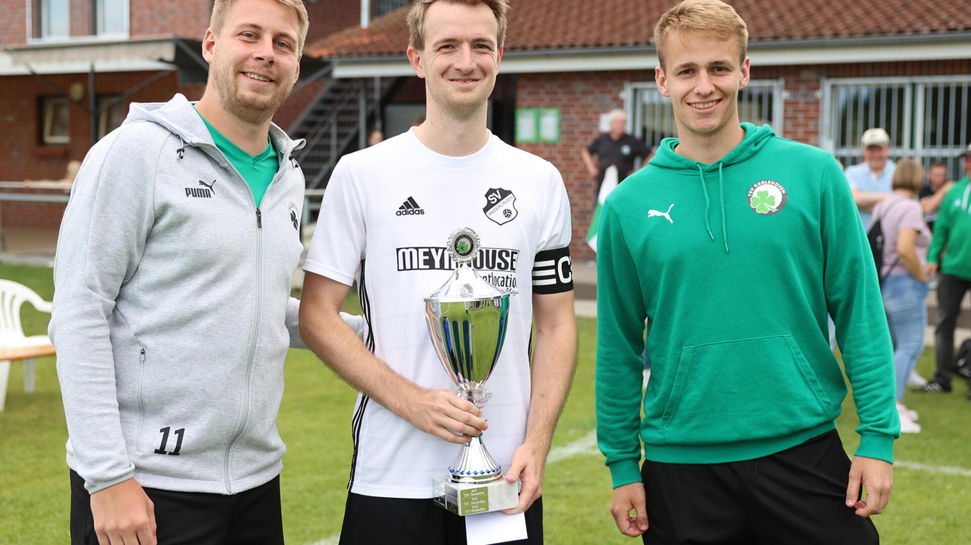 Der Favorit sicherte sich erstmalig den Titel auf der Karlshöfener Fußballwoche: Chris Bubbel (links) und Philipp Lampe (rechts) vom Karlshöfener Orga-Team gratulierten Anderlingens Kapitän Fabian Schäfer zum 3:2-Finalsieg über den FC Ostereistedt/Rhade.