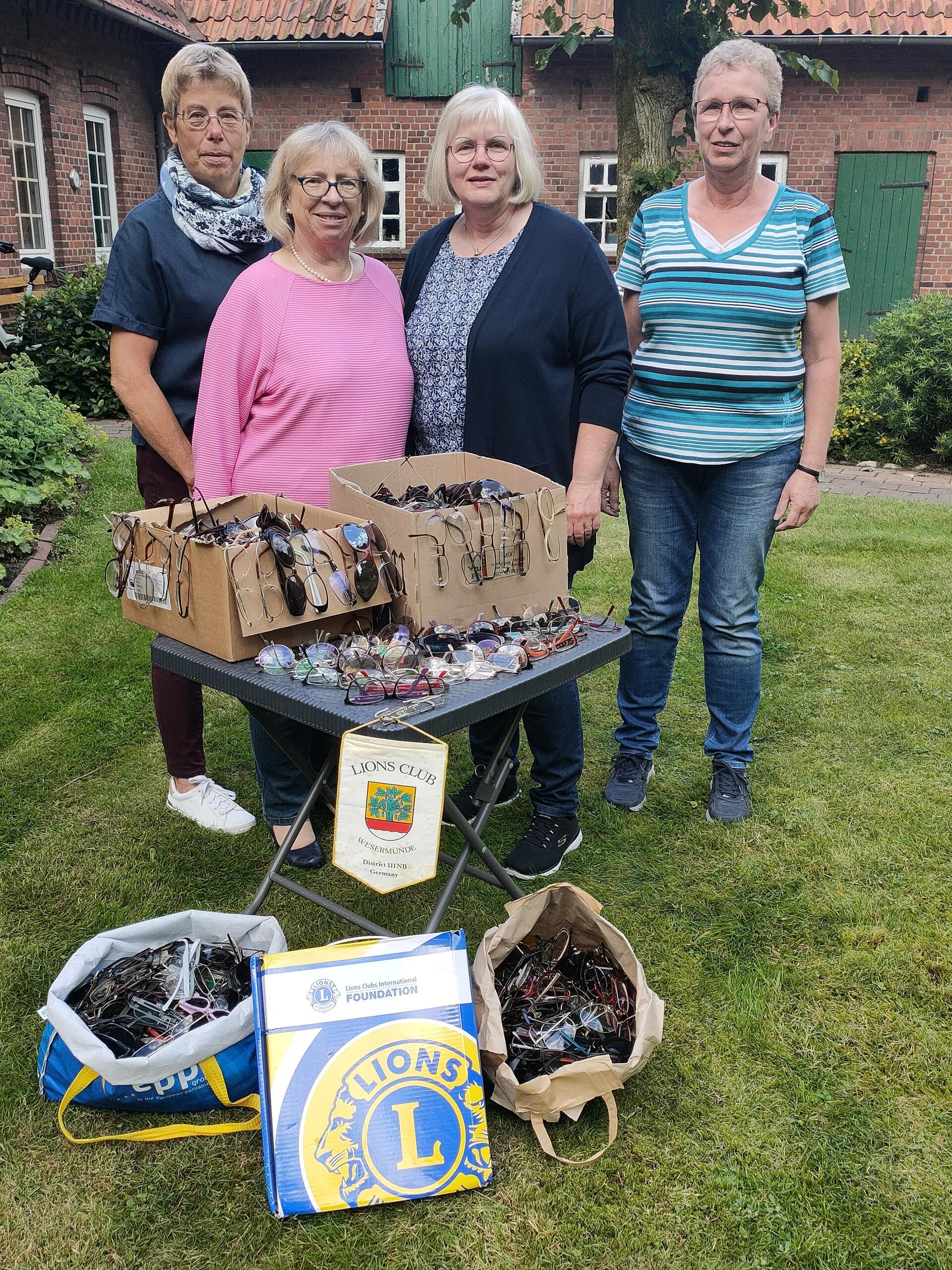 Vier Frauen stehen in einem Garten vor einem Tisch. Darauf und davor liegen Kartons und Tüten voller Brillen.