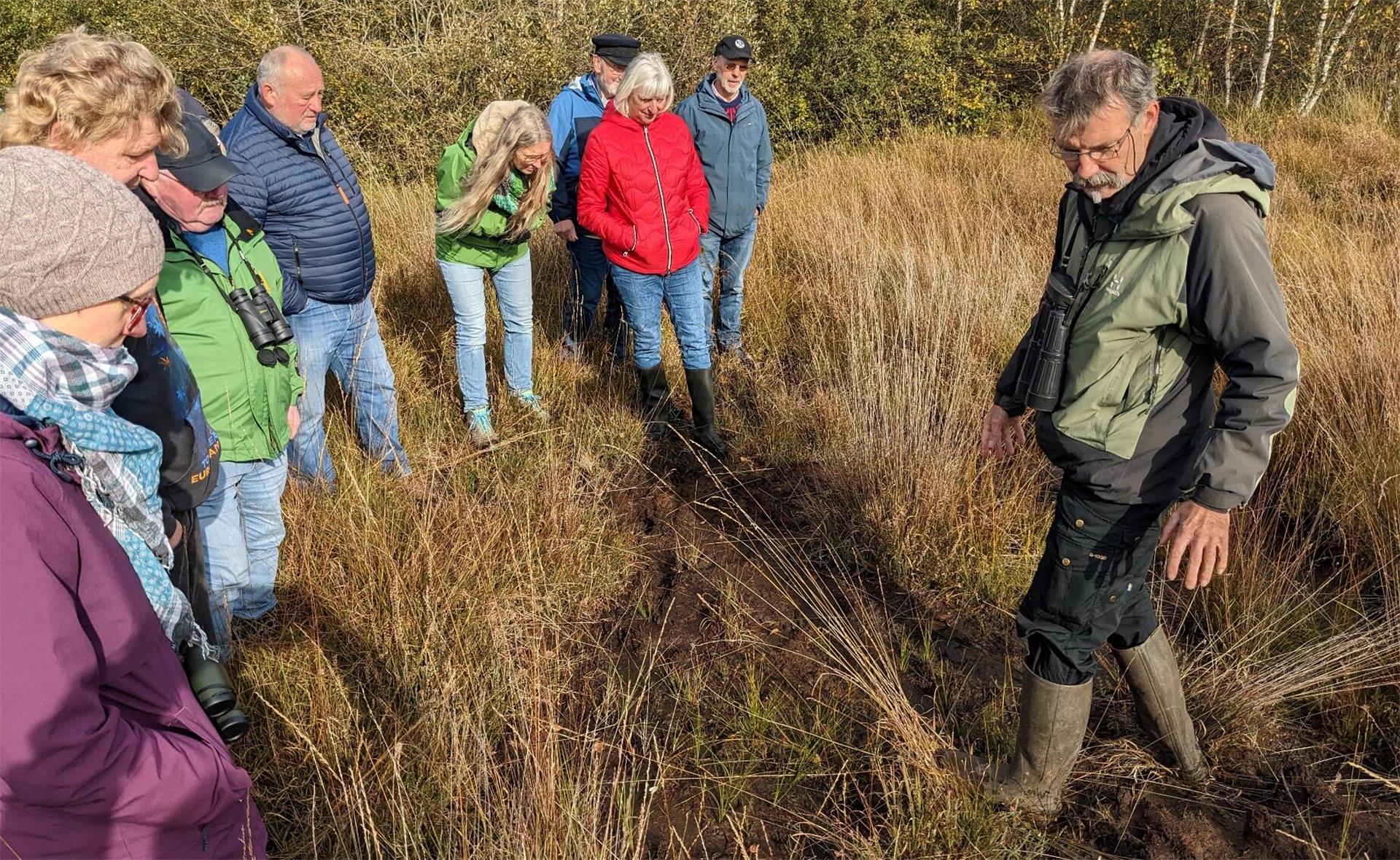 Eine Gruppe von Menschen steht in einem moorigen Gelände und hört aufmerksam einem Mann in Outdoor-Kleidung zu, der auf den Boden zeigt und scheinbar etwas erklärt. Die Personen tragen wetterfeste Kleidung und Gummistiefel. Die Umgebung ist von Gras und niedriger Vegetation geprägt.