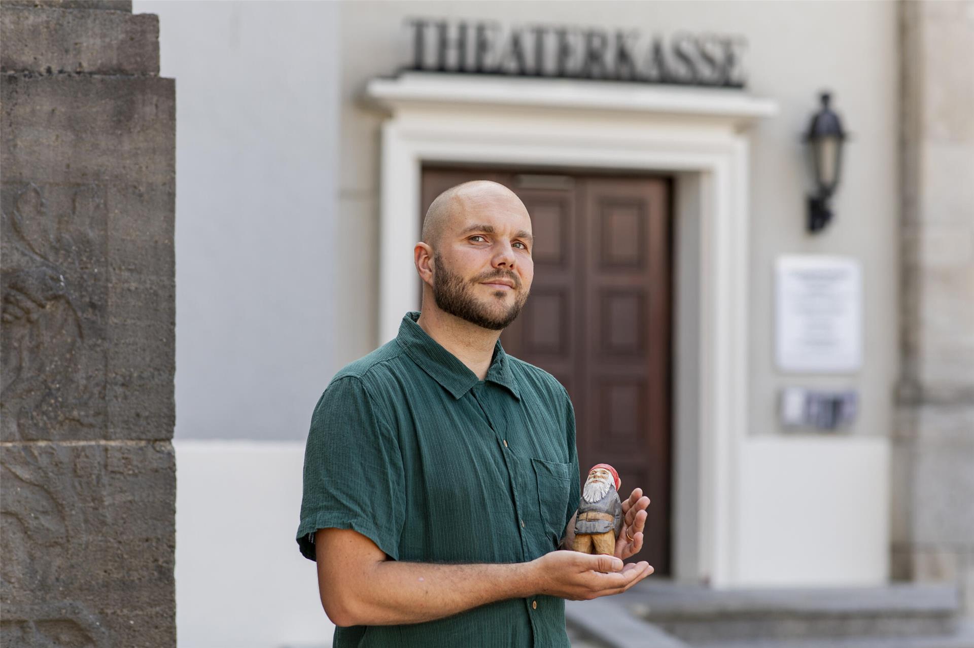 Ein Mann in einem grünen T-Shirt steht vor der Kasse des Stadttheaters. Er hält eine Holzfigur in der Hand.