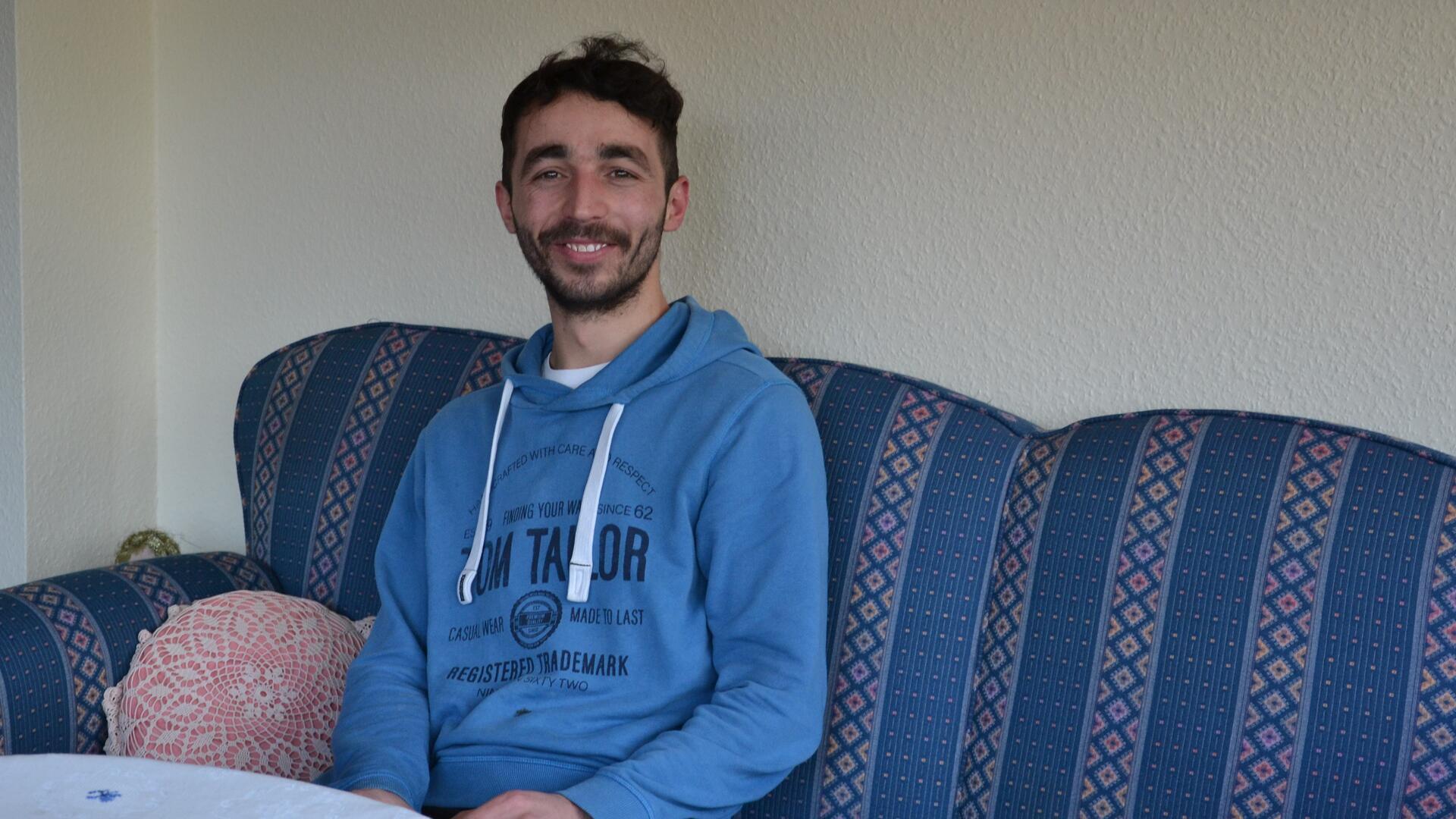 Der junge Mann aus Syrien sitzt im Wohnzimmer auf einem blauen Sofa.