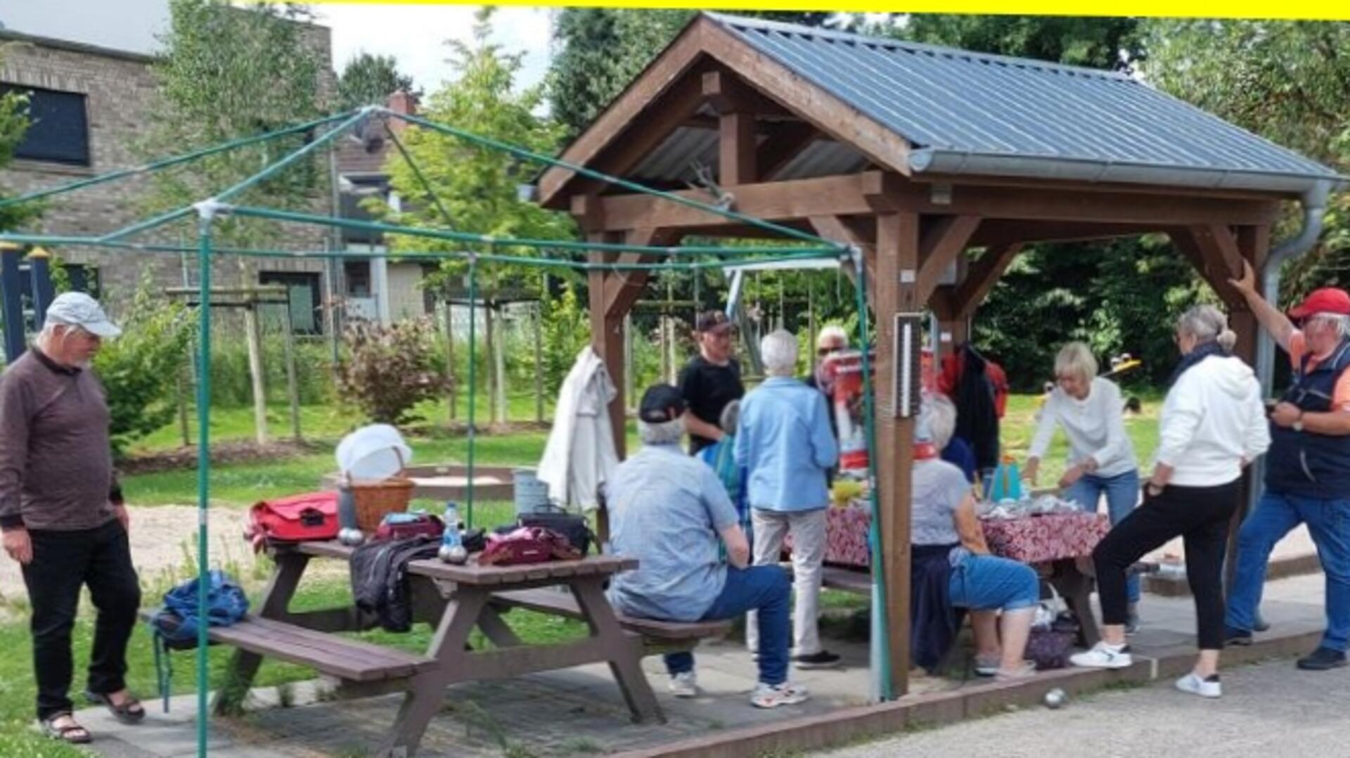 Eine Gruppe von Menschen sitzt und steht an zwei Holztischen mit Bänken. Nur einer der Tische ist überdacht.