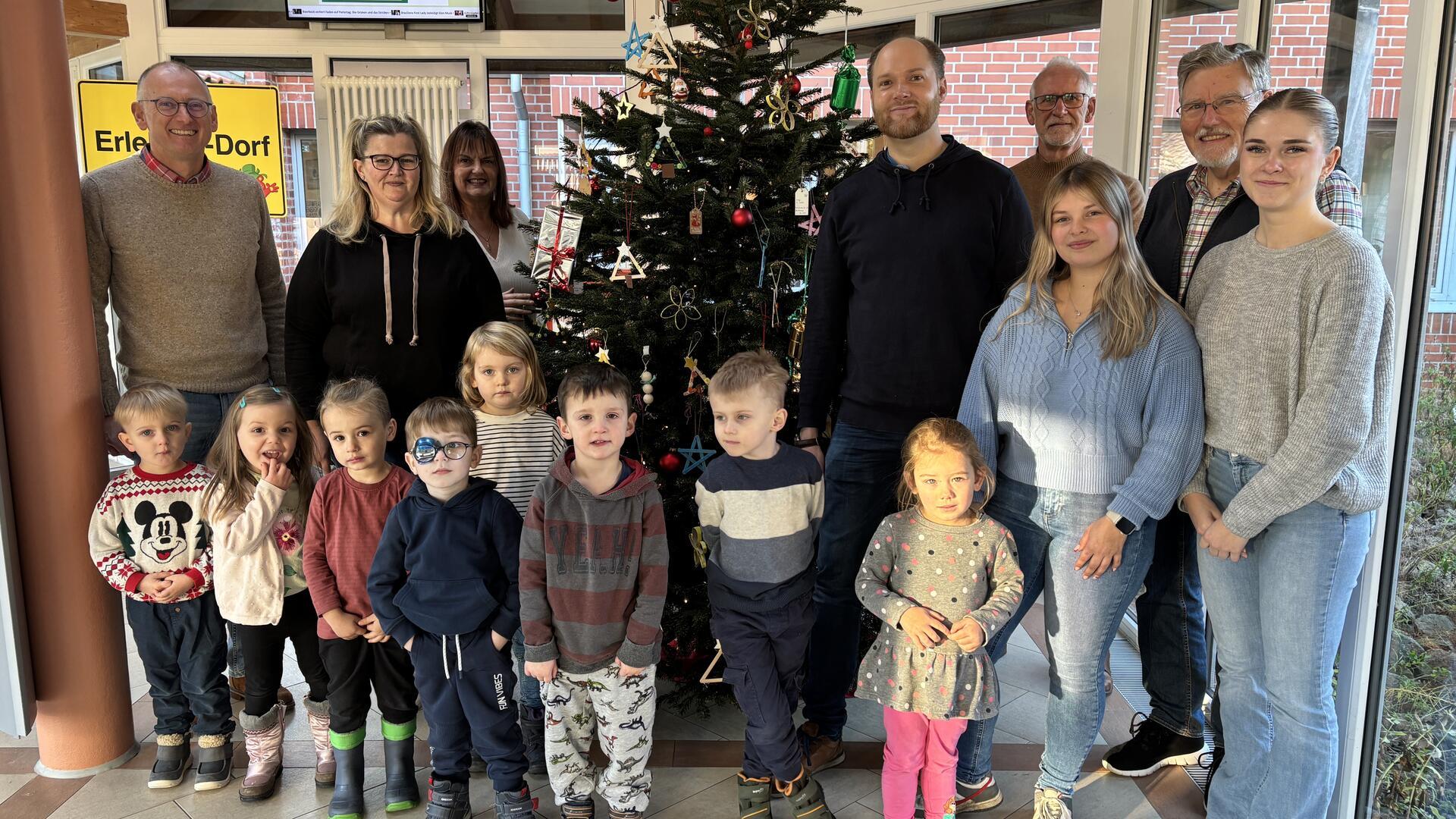 Eine Gruppe von Kindern und Erwachsenen steht vor einem weihnachtlich geschmückten Baum in einem hellen Raum mit großen Fenstern. Die Kinder tragen bunte Kleidung und Winterstiefel, einige halten die Hände verschränkt, andere schauen lächelnd in die Kamera. Im Hintergrund sind Erwachsene unterschiedlicher Altersgruppen zu sehen, die ebenfalls freundlich in die Kamera blicken. Der Weihnachtsbaum ist mit roten Kugeln, goldenen Sternen und selbst gebastelten Anhängern dekoriert. 