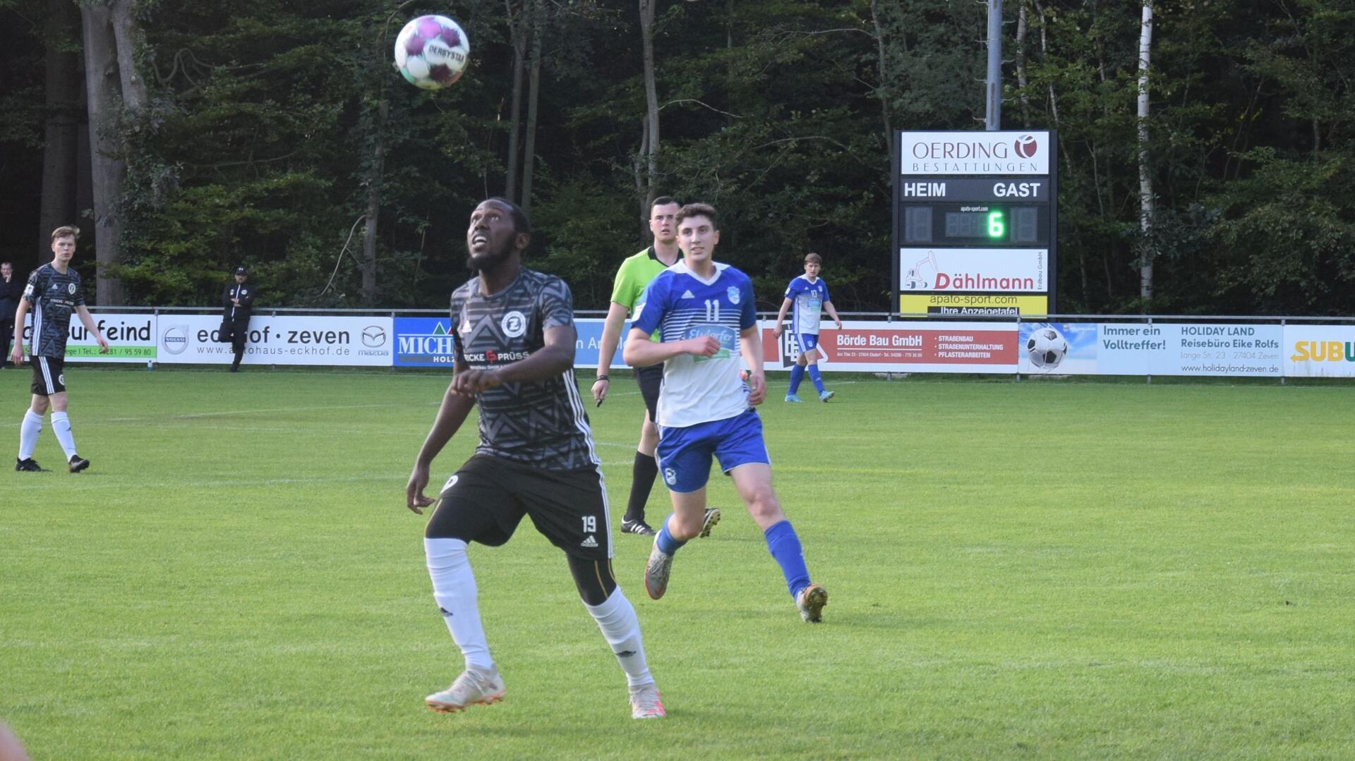 Zwei Männer in einem schwarzen und einem blauweißen Trikot laufen hinter einem Ball her.