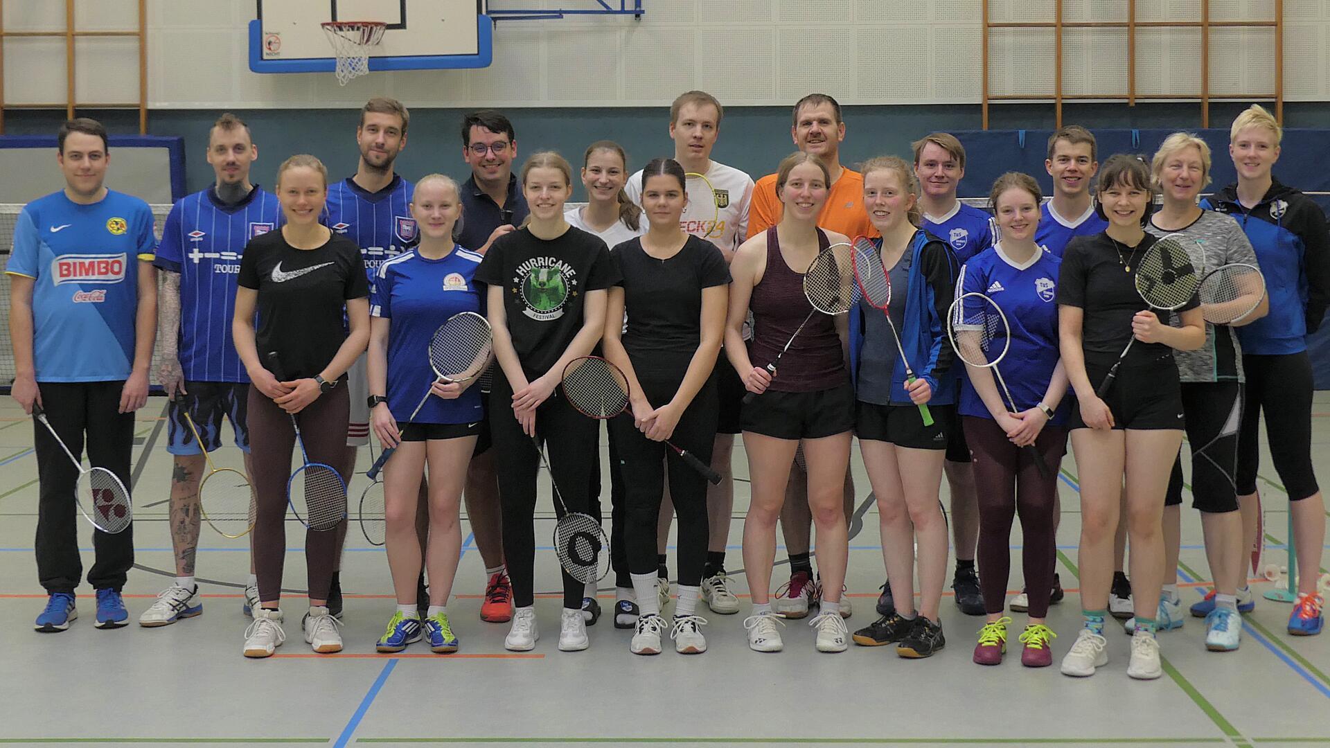 Eine Gruppe mit Sportklamotten bekleidet und Badmintonschläger in den Händen steht in einer Turnhalle. Alle lächeln in die Kamera. 