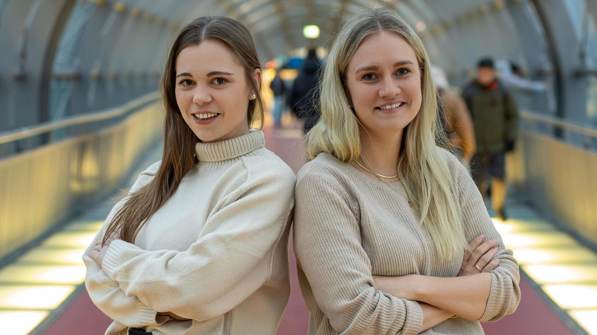 Zwei junge Frauen stehen in einem modernen, hell beleuchteten Tunnel nebeneinander und lächeln selbstbewusst in die Kamera. Beide tragen beige Pullover und haben die Arme verschränkt. 