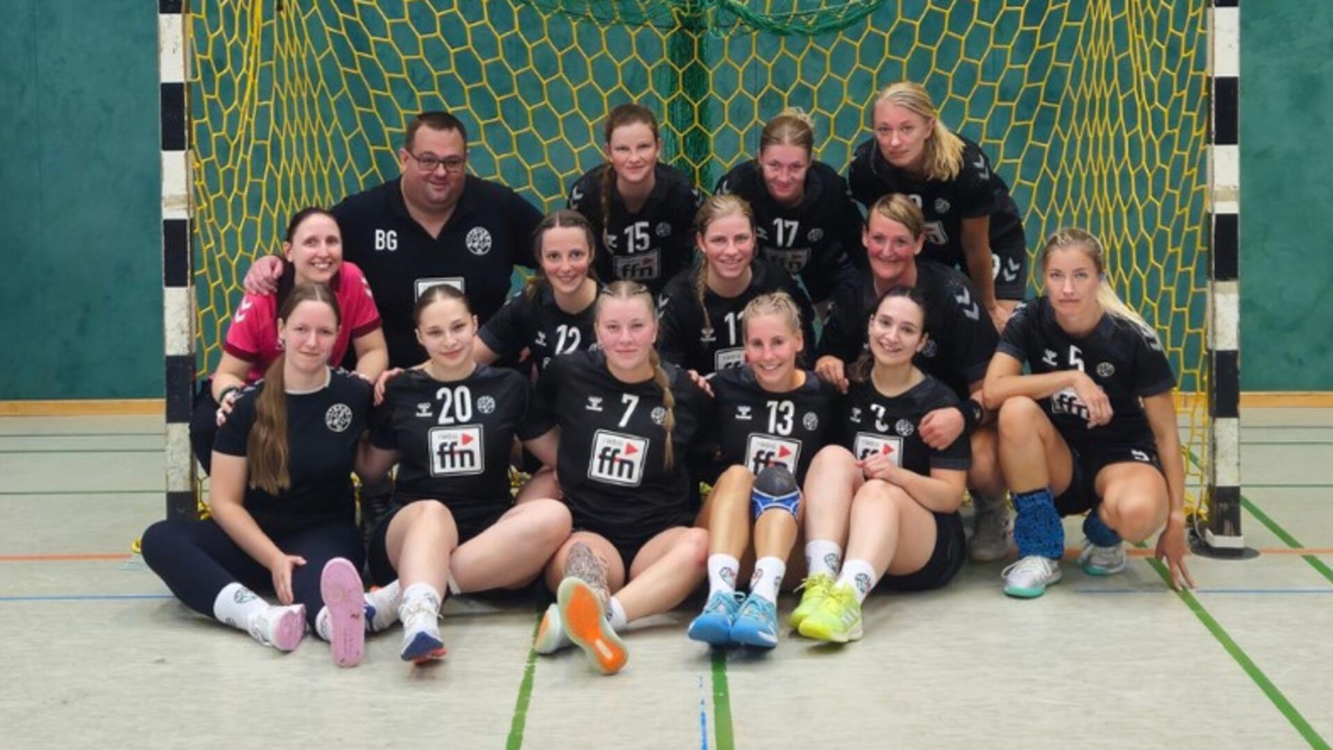 Dreizehn Frauen und ein Mann sitzen und hocken vor einem Tor in einer Sporthalle.