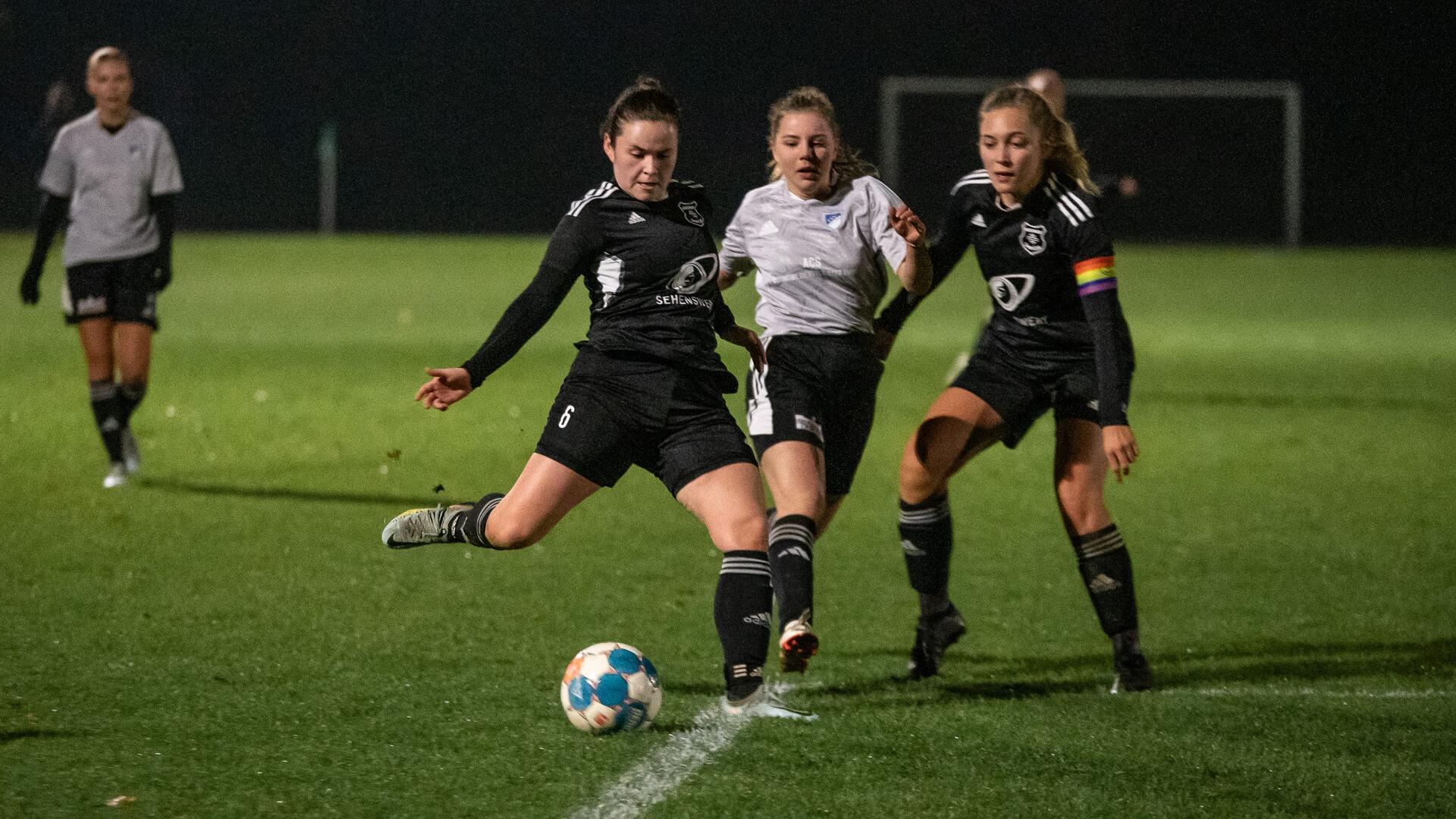 Drei Frauen in Sporttrikots laufen um einen Fußball herum.