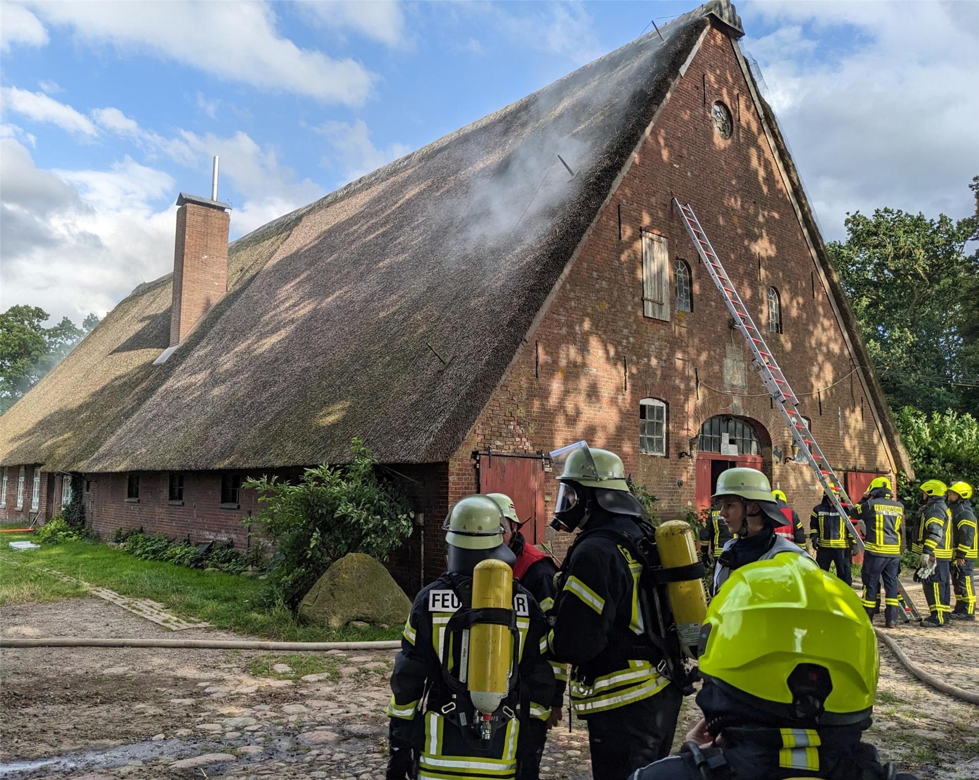 Mehrere Gruppen von Feuerwehrleuten vor einem Reetdachhaus. Eine lange Leiter lehnt an der Wand des Gebäudes.