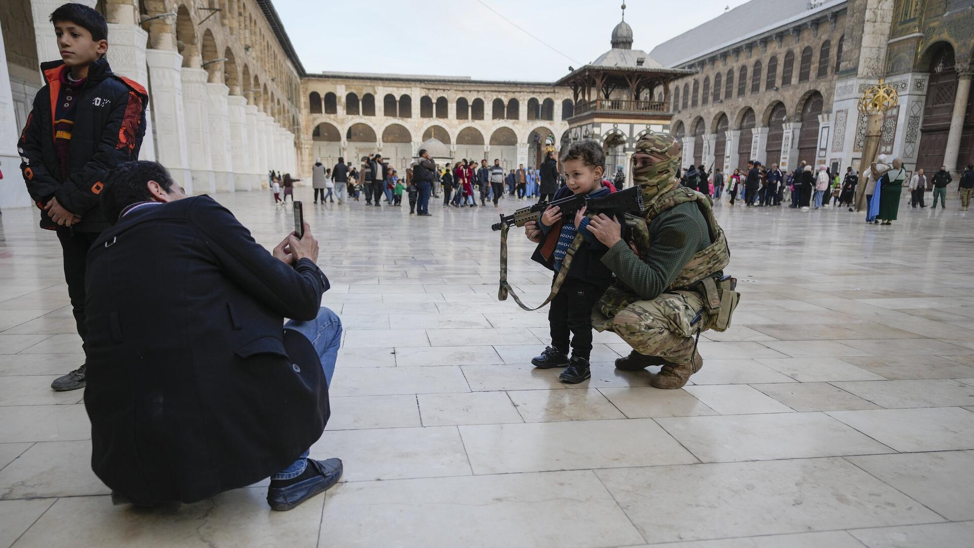 Ein Mann hockt auf einem Platz, der von Gebäuden umgeben ist und macht ein Foto von einem kleinen Jungen mit einem Gewehr in der Hand und einem Maskierten in einer Tarnuniform.