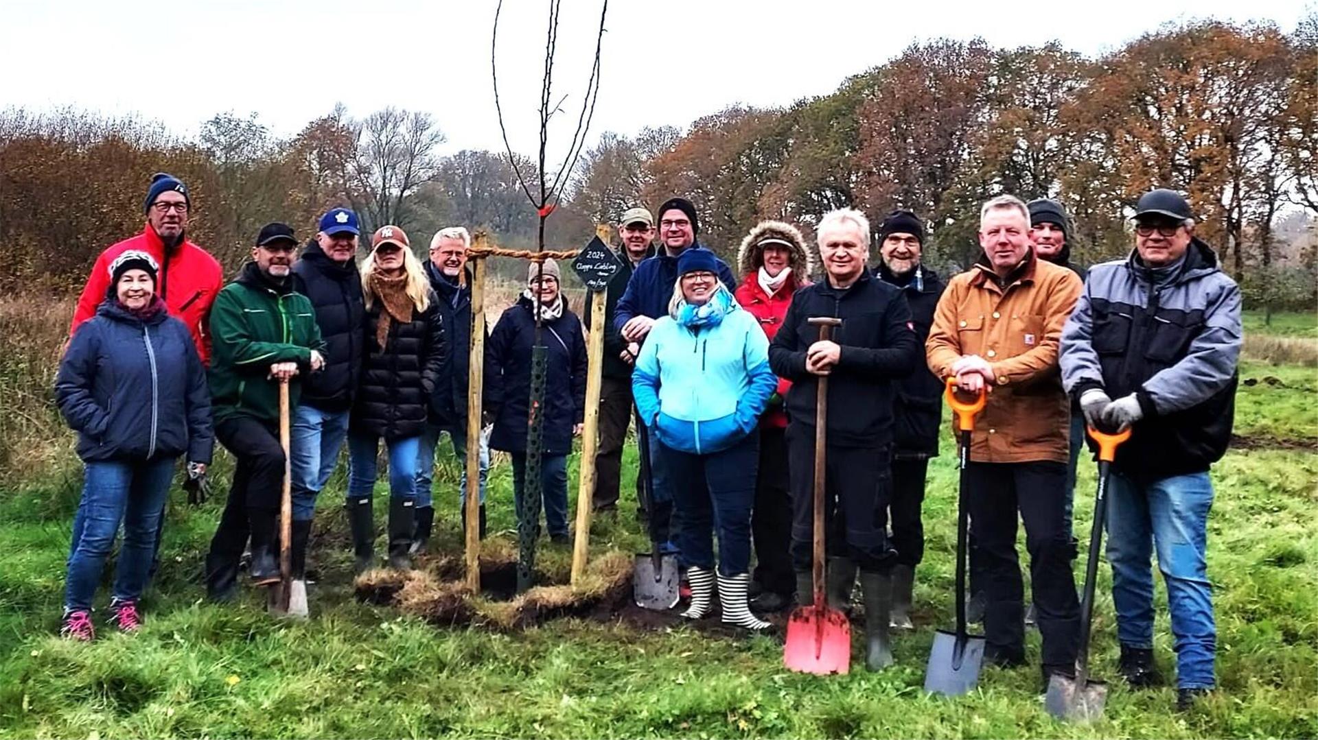 Eine Gruppe von etwa 15 Personen steht auf einer Wiese um einen frisch gepflanzten jungen Baum. Viele der Personen halten Spaten in den Händen und tragen wetterfeste Kleidung. Im Hintergrund sind herbstliche Bäume und ein bewölkter Himmel zu sehen.