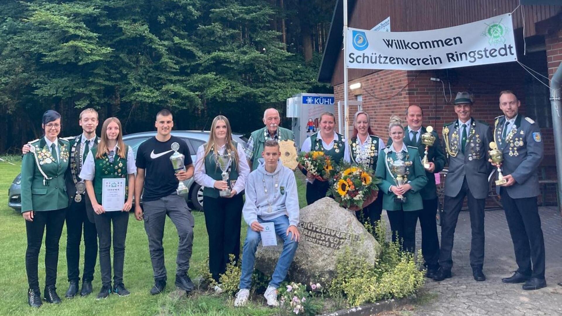 Dreizehn Schützinnen und Schützen stehen mit Urkunden und Pokalen nebeneinander. Im Hintergrund steht ein Gebäude mit einem Banner "Willkommen – Schützenverein Ringstedt"