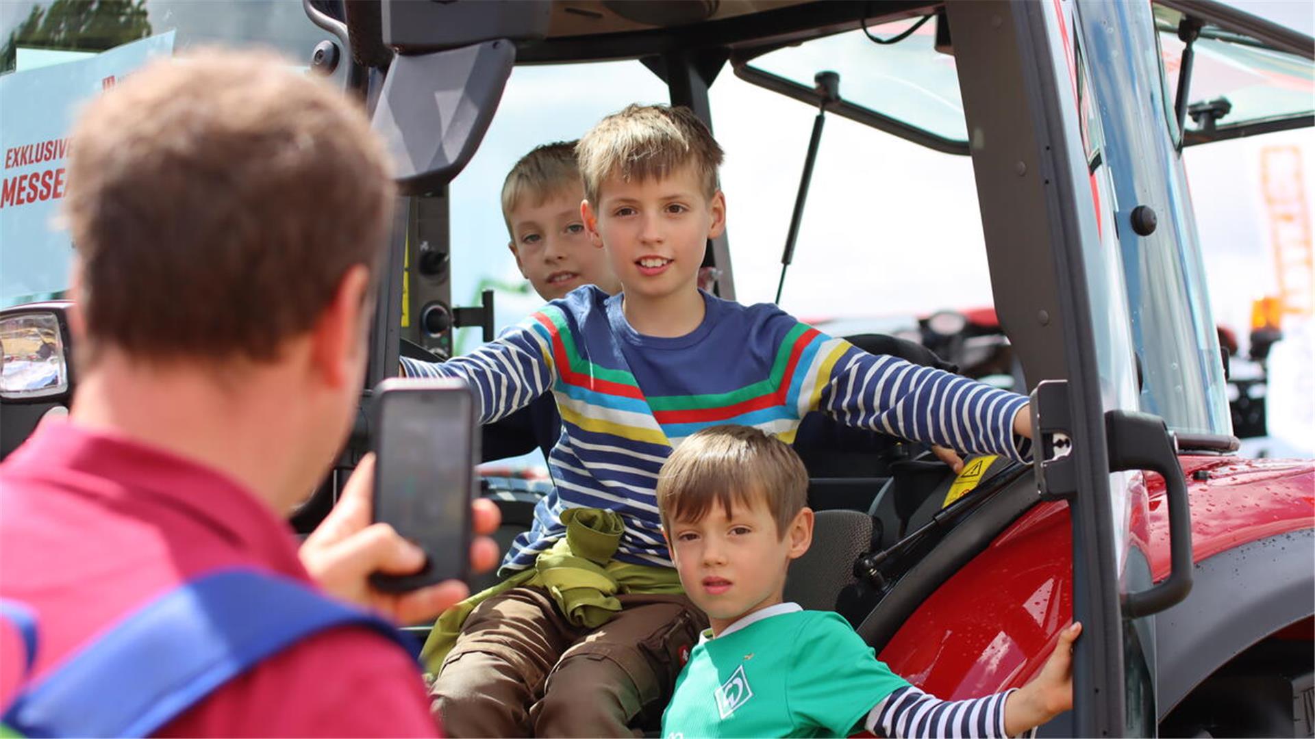 Drei Kinder sitzen auf einem Traktor und werden von ihrem Vater mit dem Smartphone fotografiert.