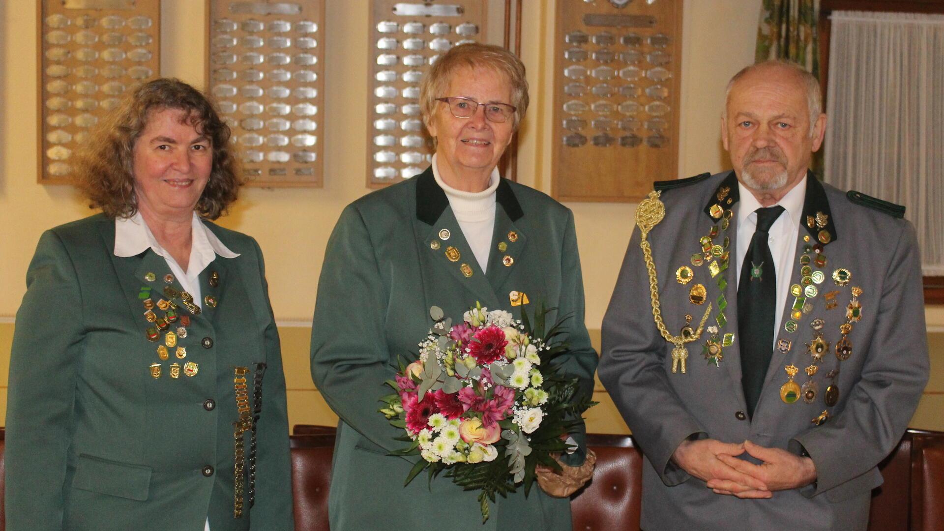 Gruppenfoto mit drei Personen in grüner und grauer Schützenuniform, dekoriert mit Abzeichen. Die Frau in der Mitte hält einen Blumenstrauß, im Hintergrund sind Ehrentafeln an der Wand zu sehen.