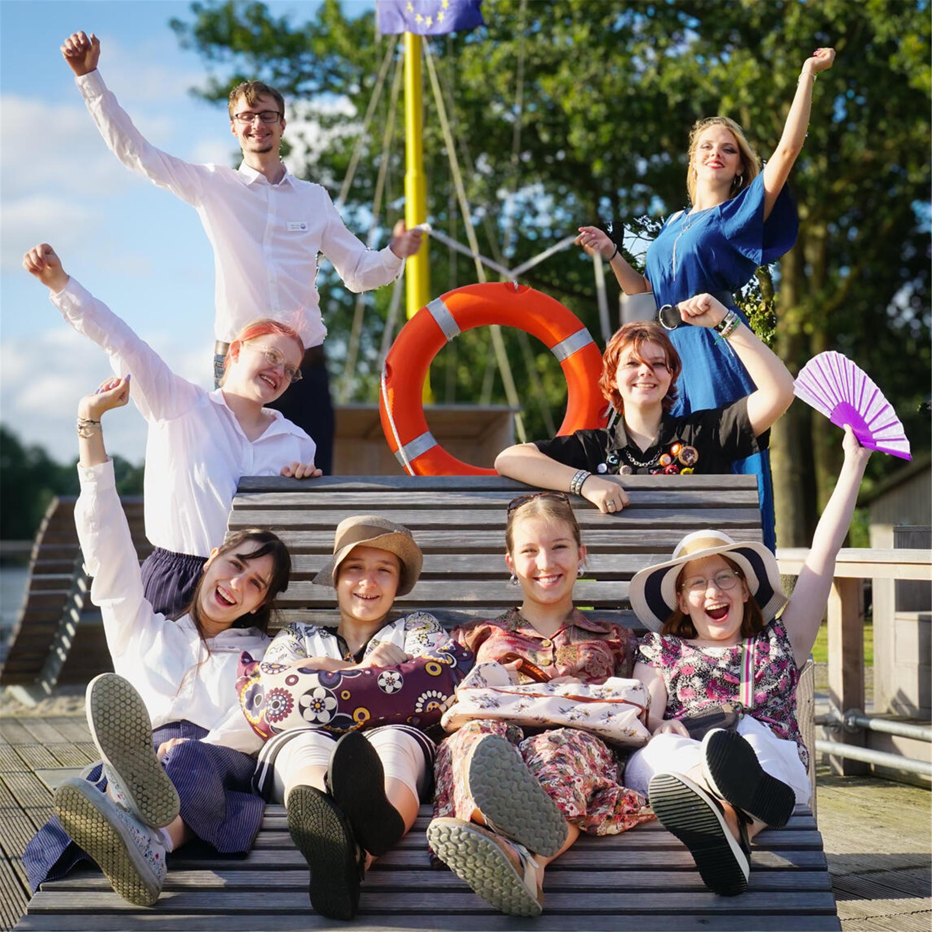 Sieben jungen Frauen und ein junger Mann sitzen und stehen rund um eine Sitzbank am Wasser. Sie lächeln und jubeln in die Kamera.