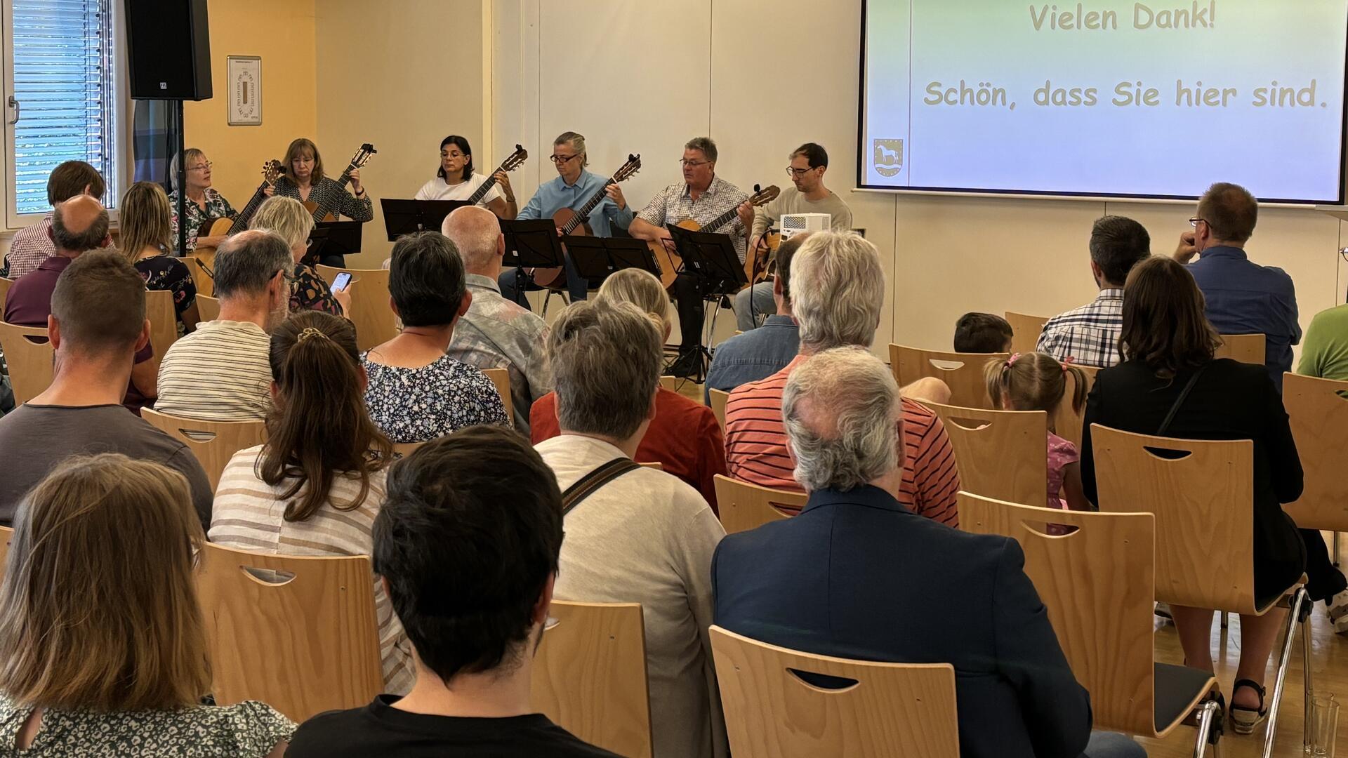 Ein Gitarrenensemble spielt vor Publikum auf Holzstühlen.