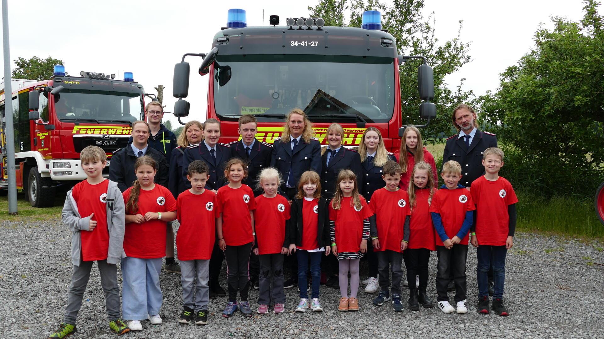 Elf Kinder in roten T-Shirts stehen in einer Reihe, hinter ihnen stehen Ältere und Erwachsene, größtenteils in Feuerwehruniform. Im Hintergrund stehen zwei Feuerwehrfahrzeuge.