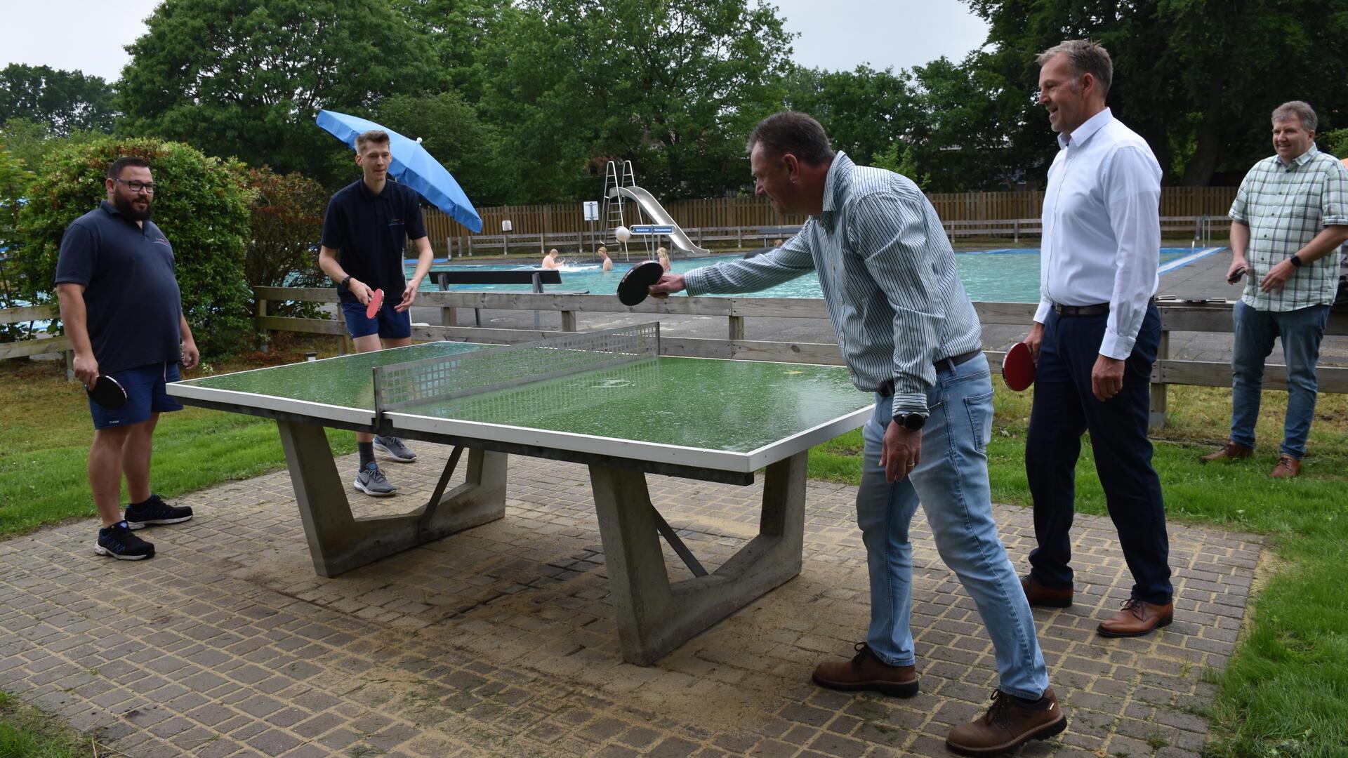 Fünf Männer spielen an einer Tischtennisplatte im Freien, im Hintergrund schwimmen Menschen in einem Freibad.