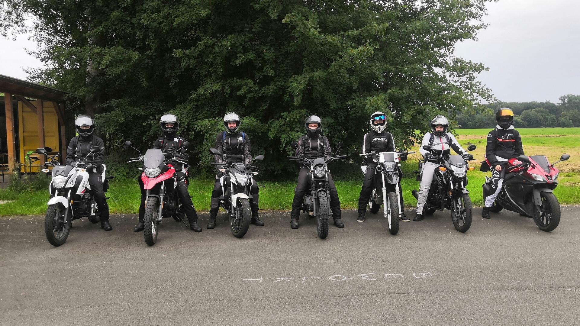 Zu sehen sind sieben Motorradfahrer, die auf aufgereiht auf einem Parkplatz stehen. 