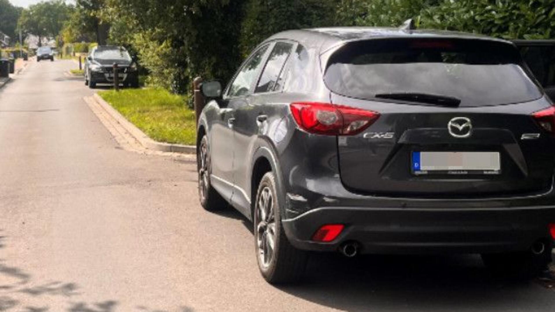 Ein schwarzes Auto steht rechts in einer Parkbucht und hat eine Schramme auf der linken Seite der hinteren Stoßstange.