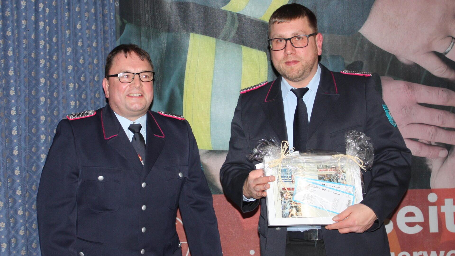 Zwei Männer in Feuerwehruniformen stehen nebeneinander und lächeln in die Kamera. Der Mann rechts hält ein Geschenkset in den Händen, das in Folie verpackt und mit einer Schleife versehen ist.