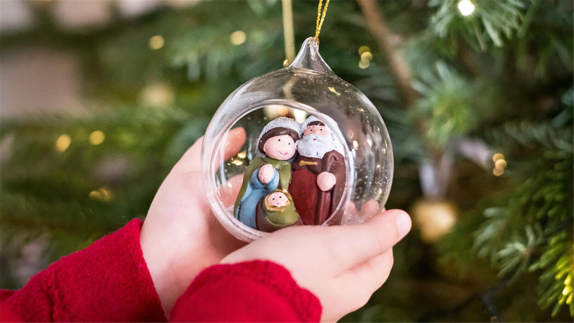 Ein Kind hält eine Krippe mit Maria, Josef und dem Jesuskind in einer gläsernen Weihnachtskugel an einem Weihnachtsbaum.