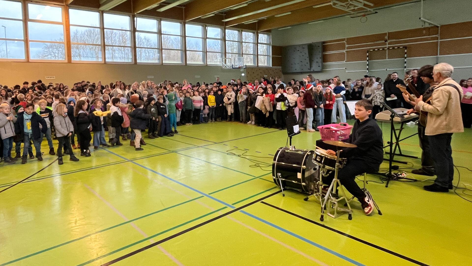 Eine große Gruppe von Kindern und einige Erwachsene versammeln sich in einer Sporthalle. Im Vordergrund ist ein Schüler an einem Schlagzeug zu sehen, begleitet von weiteren Musikern mit Gitarren und einem Keyboard. 