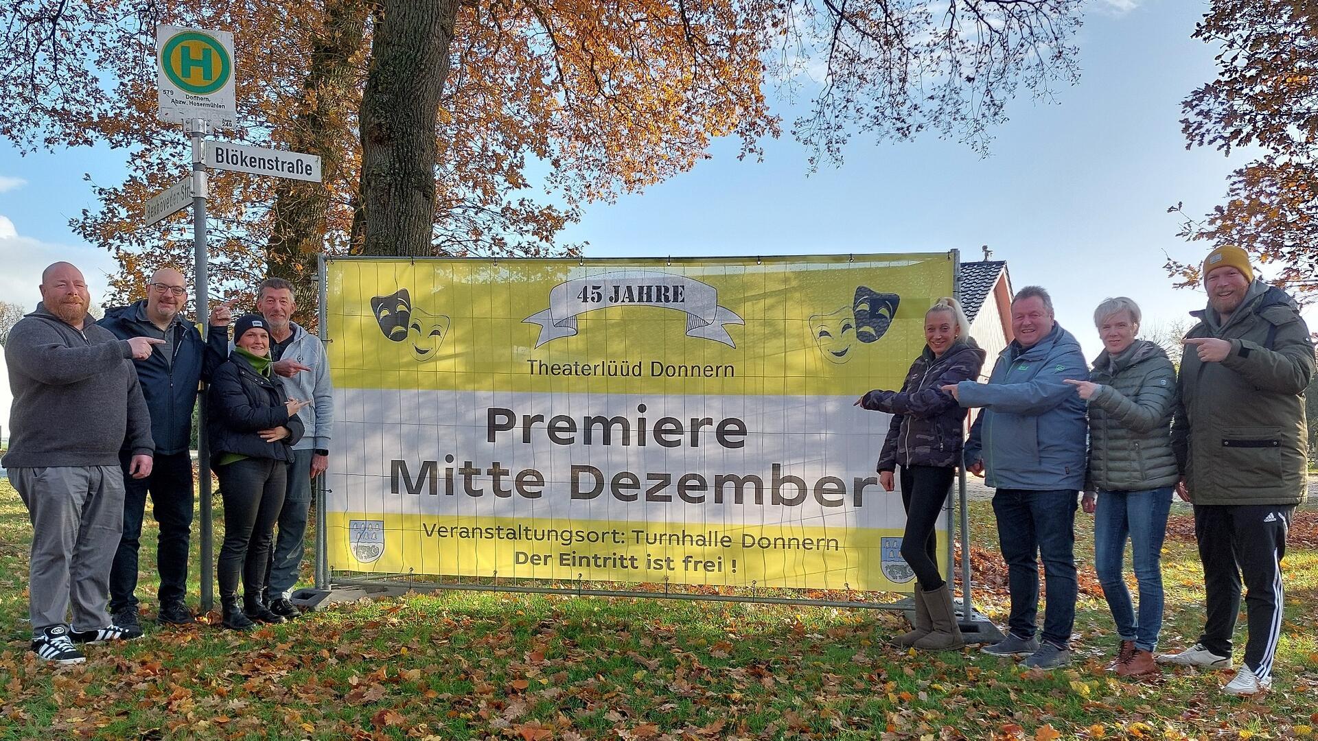 Eine Gruppe von Menschen steht in einer herbstlichen Parklandschaft vor einem großen gelben Banner. Auf dem Banner steht in schwarzer Schrift: „Premiere Mitte Dezember“. Die Menschen lächeln und zeigen auf das Banner, um die Veranstaltung anzukündigen.