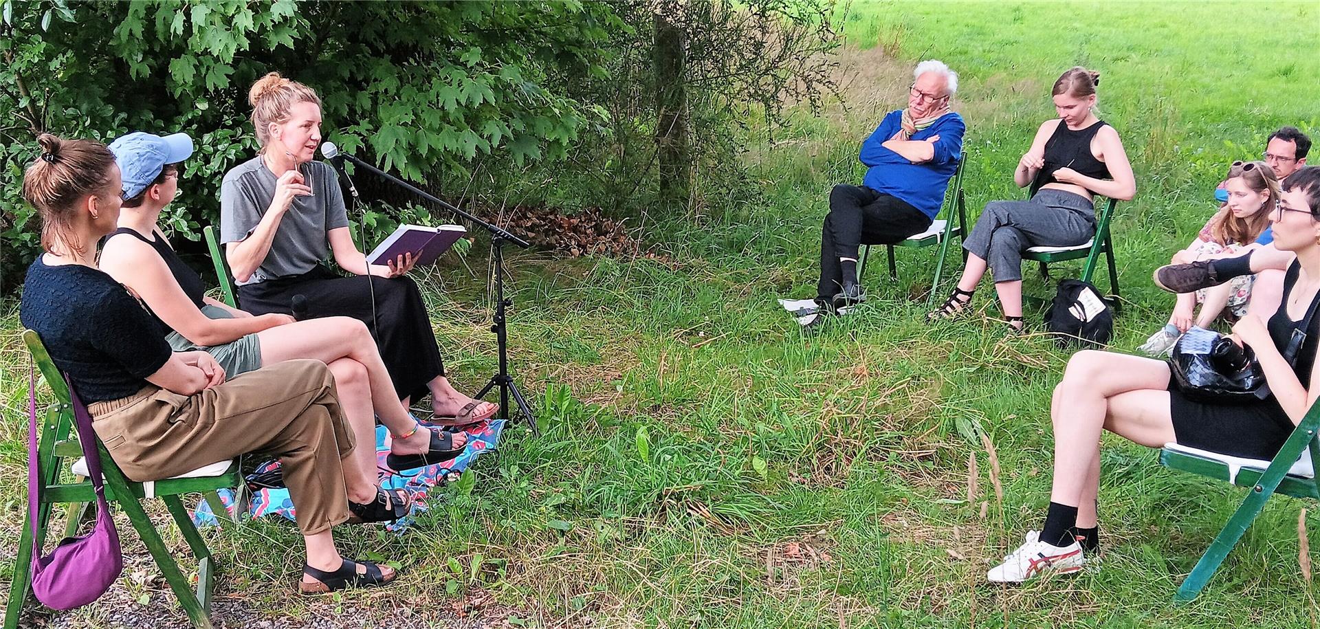 Einige Frauen und Männer sitzen in einem Kreis auf Klappstühlen auf einer Grünfläche. Eine Frau hält ein Buch und spricht in ein Mikrofon.