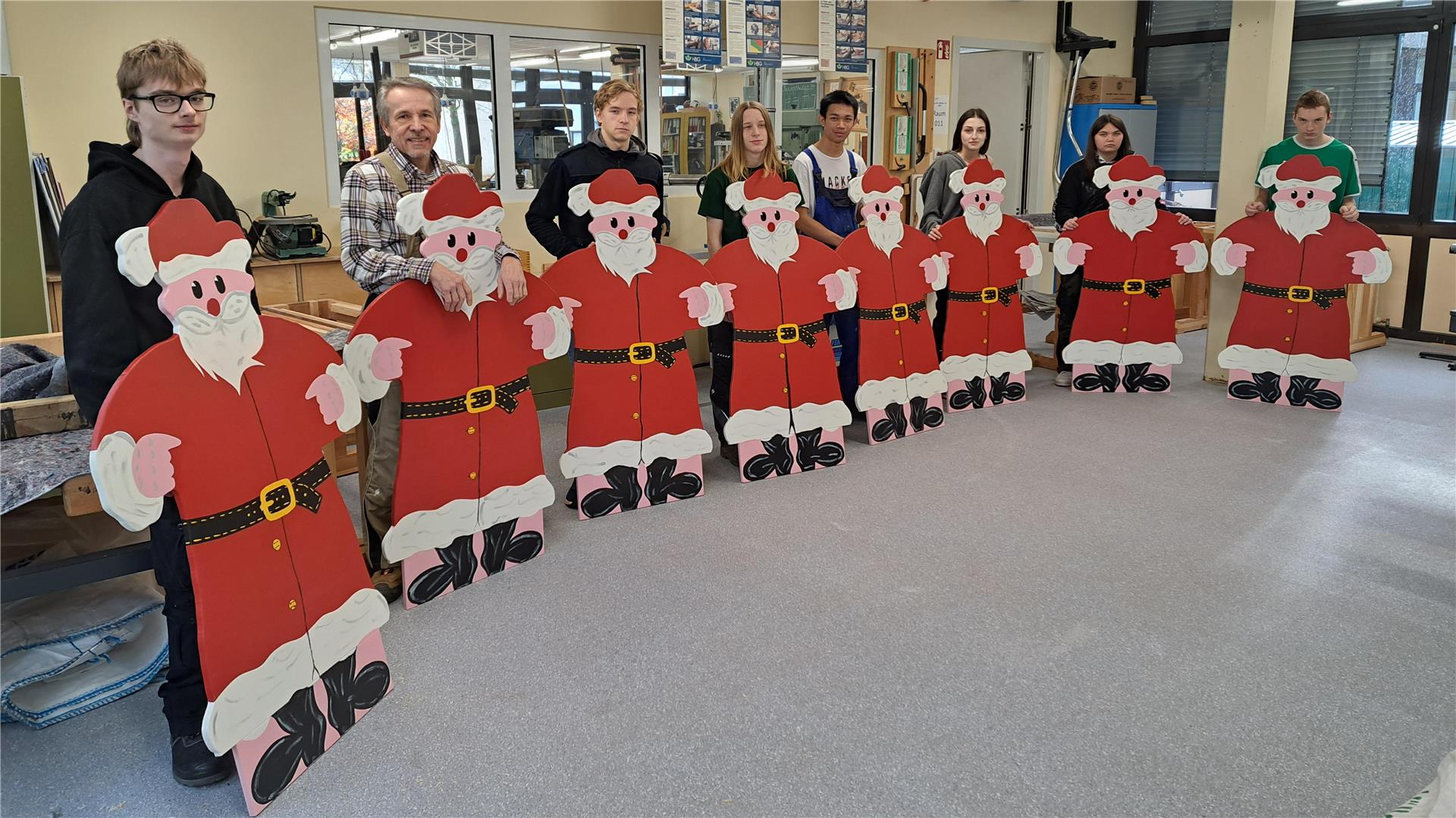 Eine Gruppe von Schülerinnen, Schülern und einem Lehrer steht in einem Werkraum und präsentiert acht große, selbst gebaute und bemalte Weihnachtsmannaufsteller. Die Figuren aus Sperrholz sind in kräftigem Rot, Weiß und Schwarz gestaltet und zeigen fröhliche Weihnachtsmänner