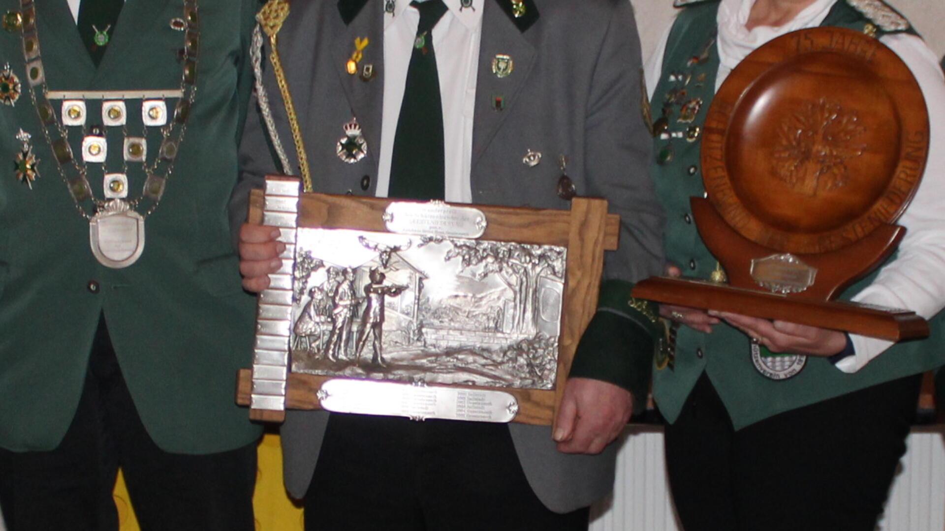 Detailaufnahme von drei Personen in Schützenuniformen. Ihre Uniformen sind mit Abzeichen und Orden geschmückt. Sie halten eine Holzplakette mit eingearbeiteter Metallgravur und eine hölzerne Ehrentafel.