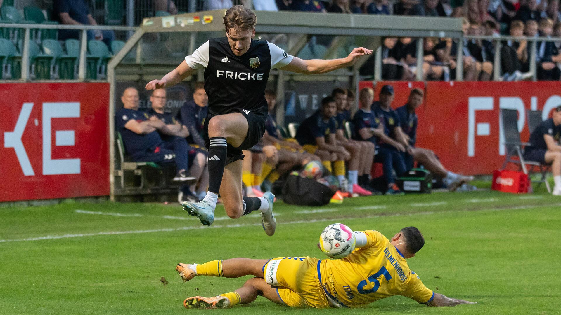 Ein Mann in einem schwarzen Trikot springt über einen liegenden Mann im gelben Trikot.