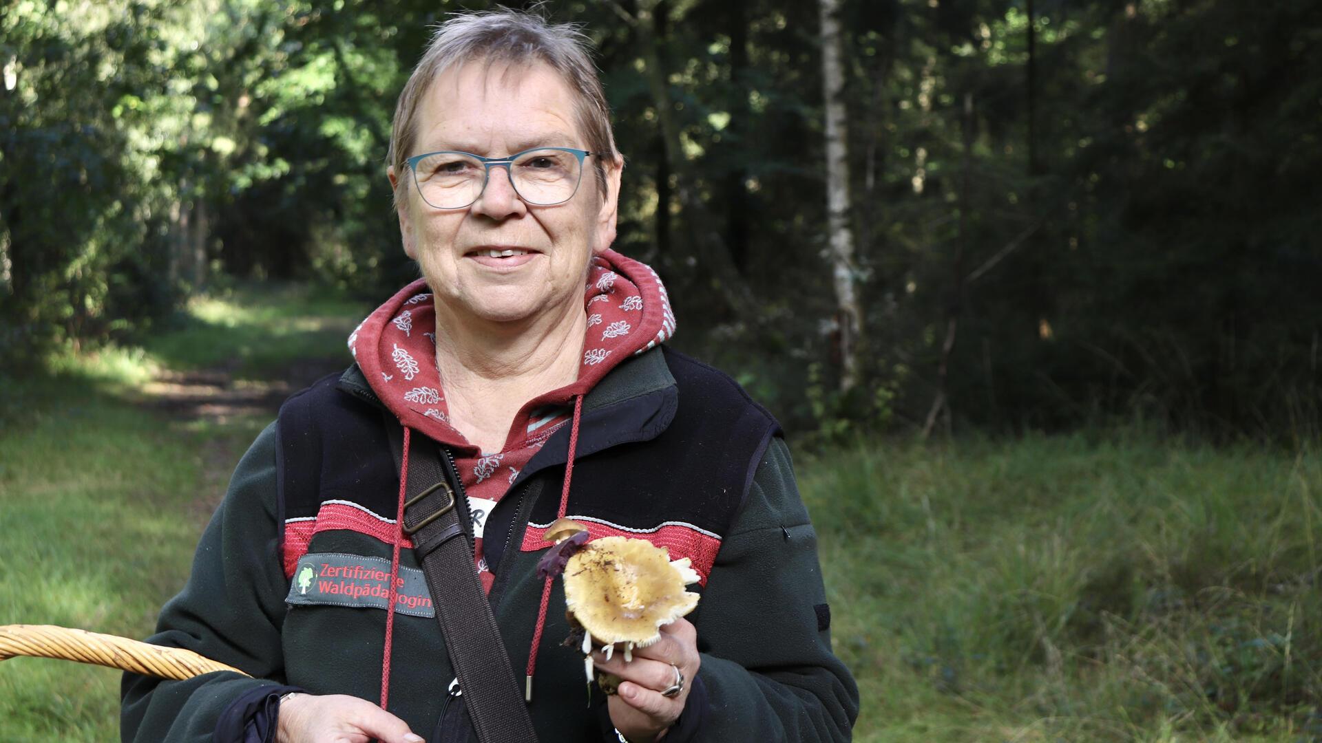 Eine Frau hält einen Pilz in der Hand und blickt in die Kamera.