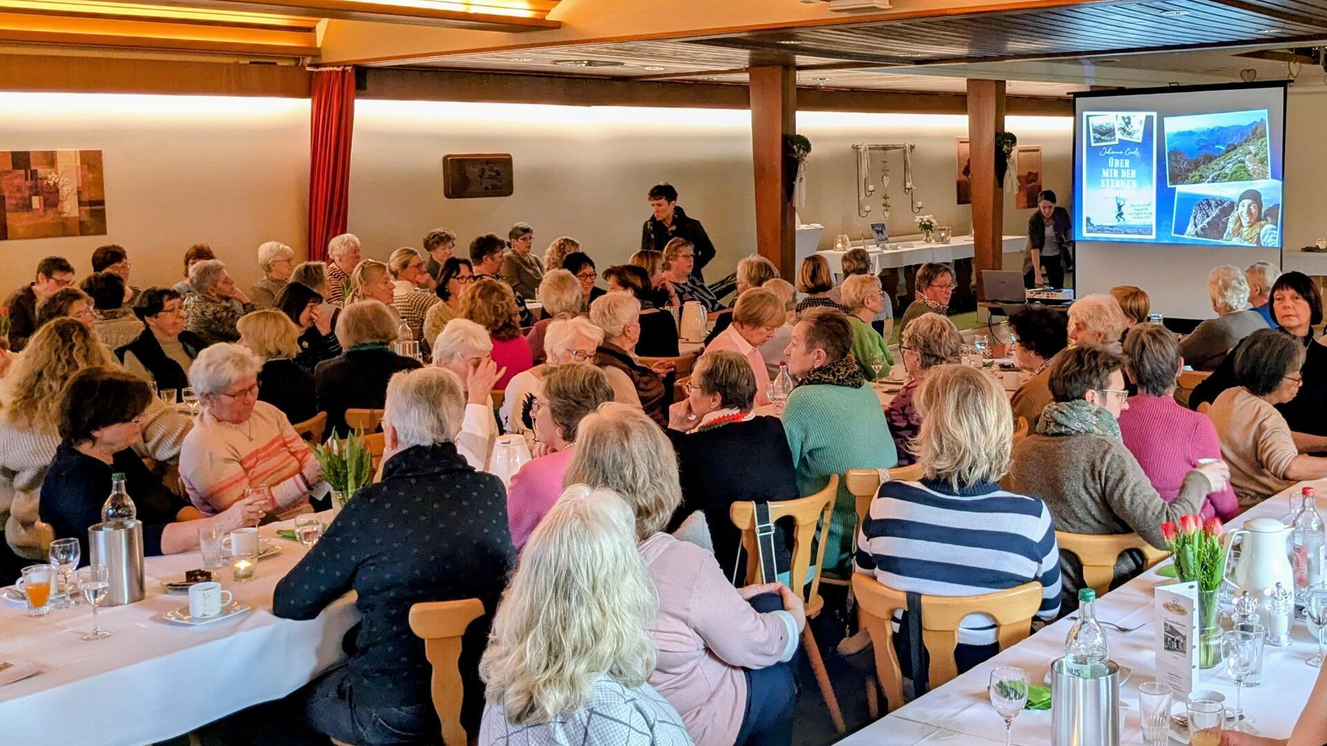 Eine Versammlung in einem Saal, in dem zahlreiche Frauen an Tischen mit weißer Tischdecke sitzen. Im Hintergrund ist eine Leinwand mit einer projizierten Präsentation zu sehen. Die Tische sind mit Getränken, Gläsern, Kaffeetassen und Dekorationen wie Blumen geschmückt.