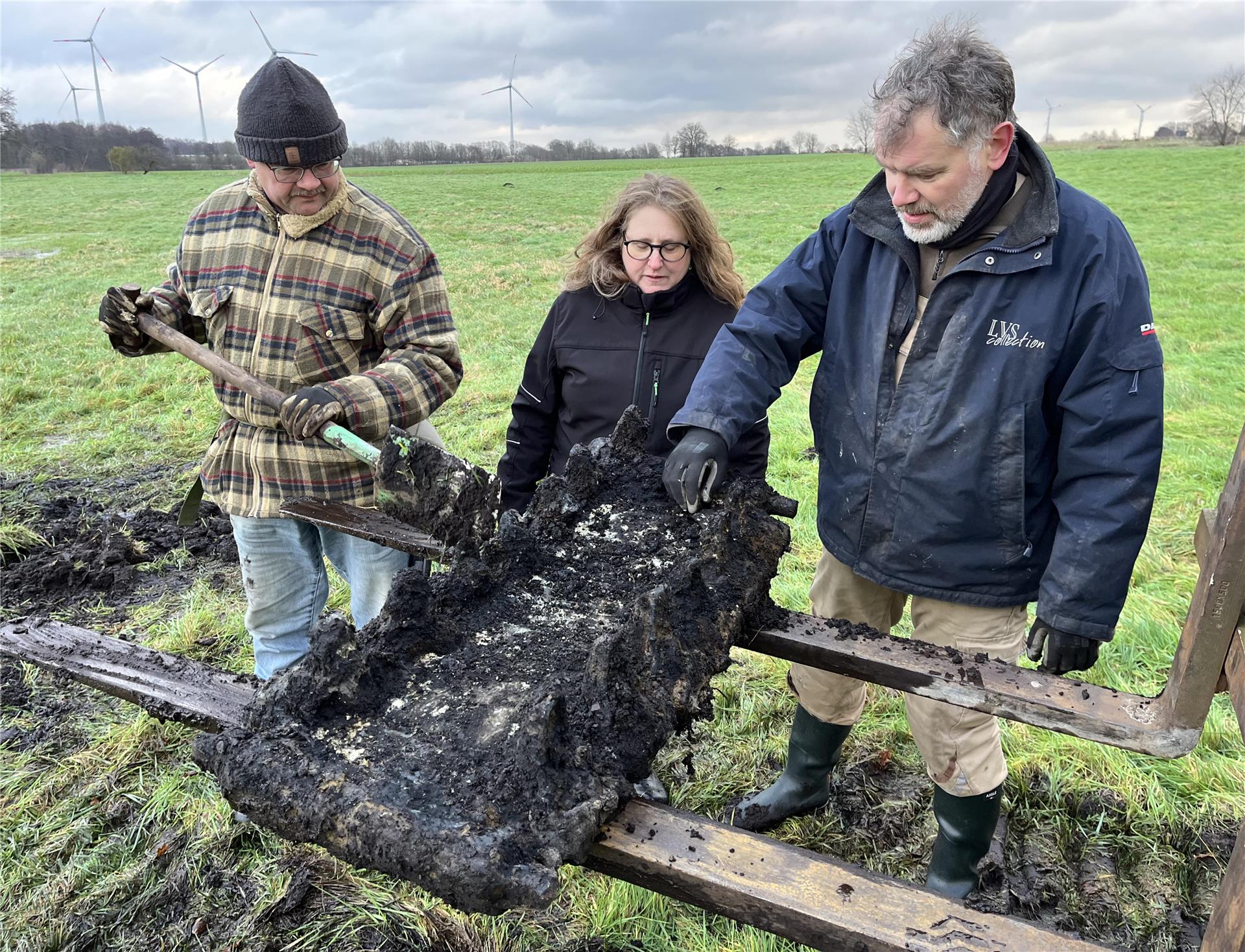 Frank Hoferichter, Debbie Bülau und Kreisarchäologe Daniel Nösler betrachten das Fragment der Panzerkette eines britischen Sherman-Panzers auf einer Wiese bei Kutenholz (von links).