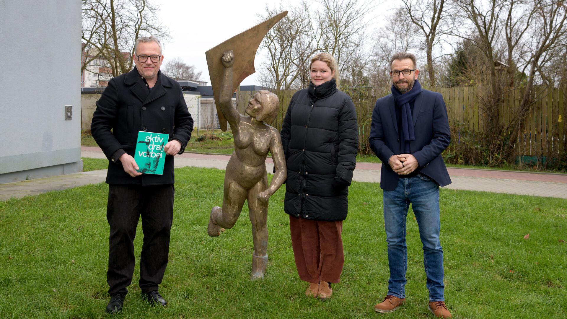 Drei Personen stehen auf einer Grünfläche vor einer lebensgroßen Bronzeskulptur, die eine Frau beim Drachensteigen darstellt. 