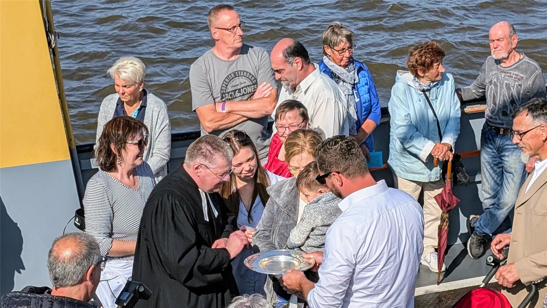 Menschengruppe an Bord einer Fähre auf dem Wasser. Ein Pastor steht im Kreis mit einer Familie und tauft ein Kind.