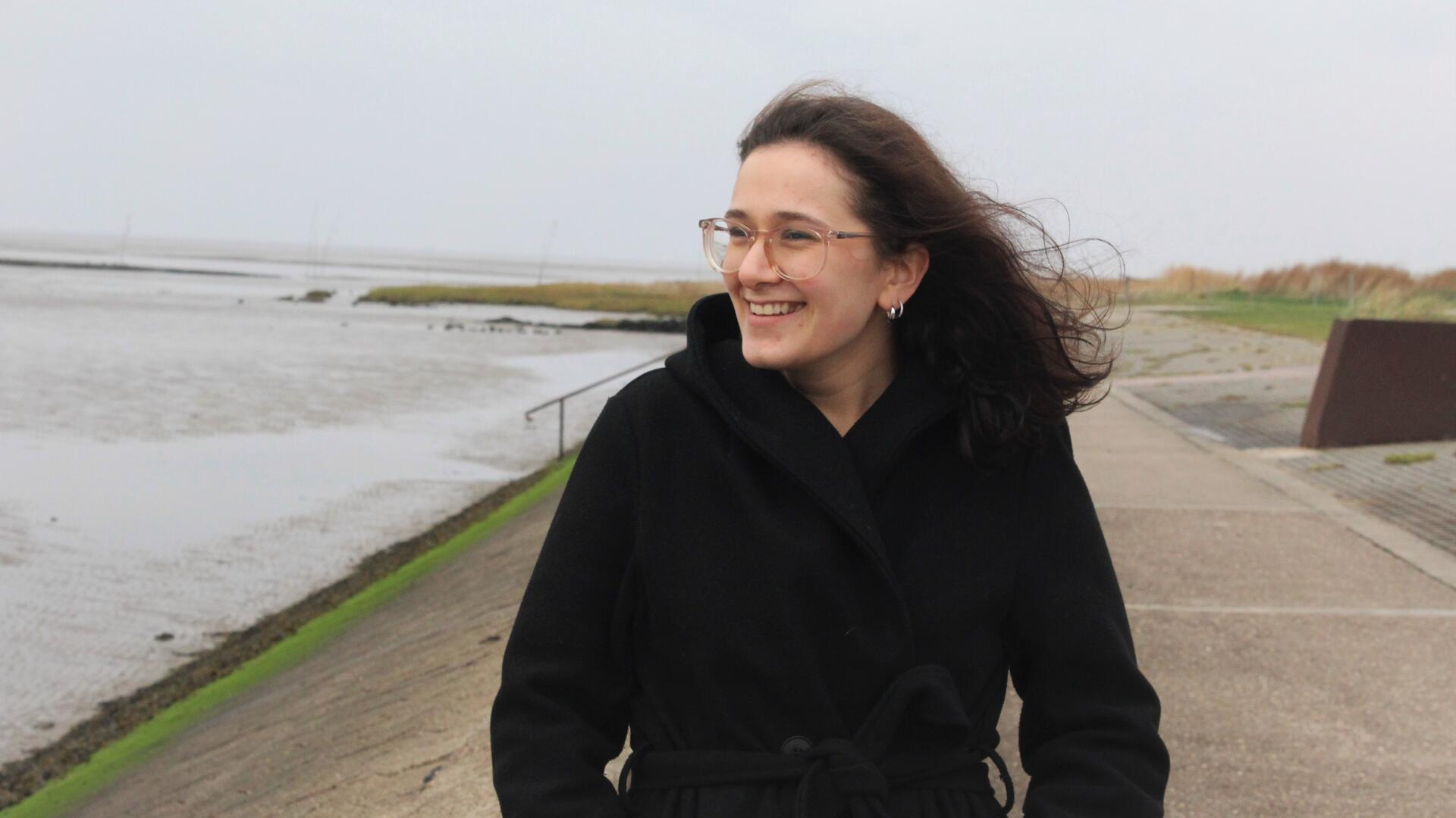 Frau mit schwarzer Jacke steht am Strand.