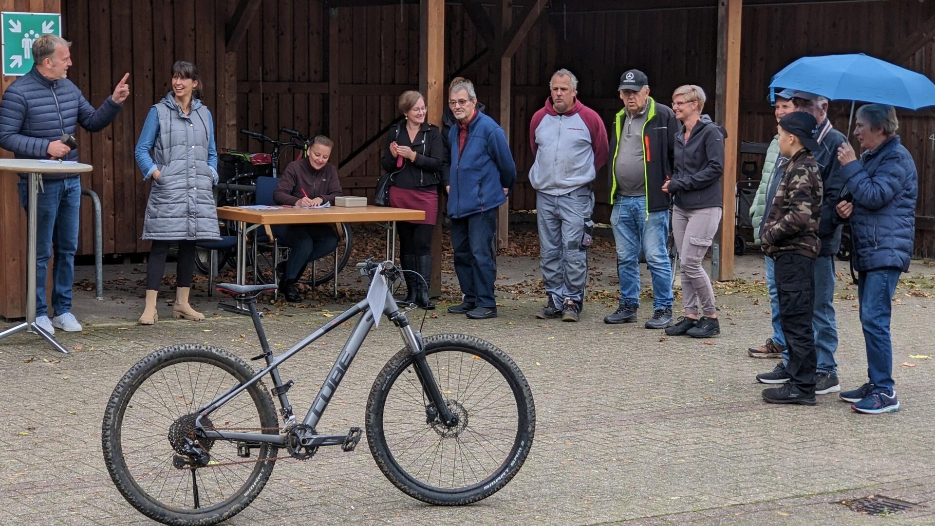 Auf einem Parkplatz steht eine Gruppe von Menschen um ein silbergraues Mountainbike herum.