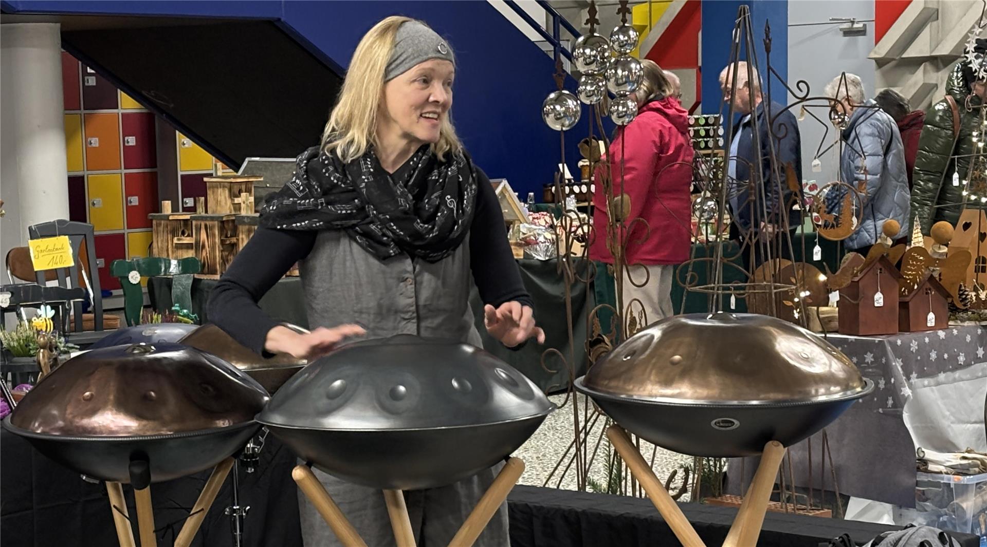 Eine Frau spielt auf mehreren Handpans, die auf Ständern nebeneinander aufgereiht sind. Sie trägt ein Stirnband und einen Schal, während sie das Instrument in einer Ausstellungsumgebung präsentiert.