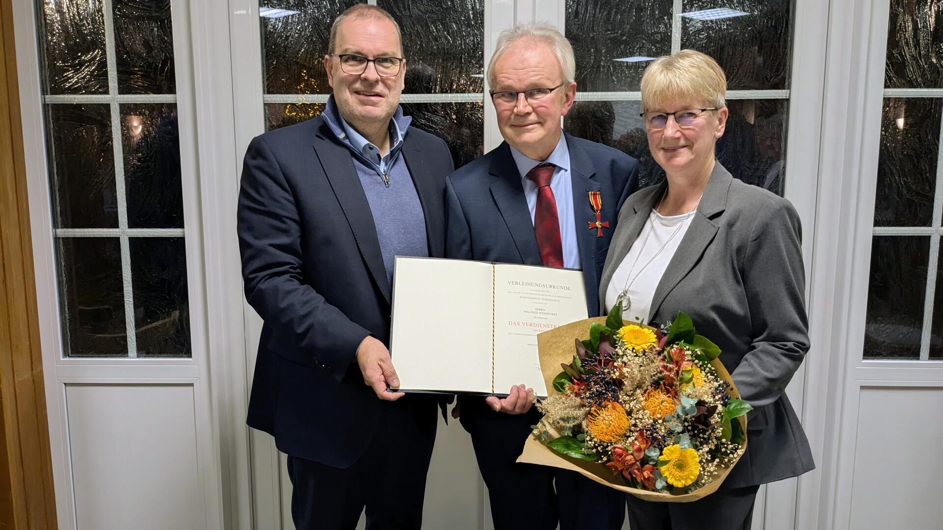 Drei Personen stehen nebeneinander vor einer Fenstertür mit weißen Rahmen. Der Mann in der Mitte hält eine Verleihungsurkunde in der Hand und trägt ein Bundesverdienstkreuz am Anzug. Die Frau rechts hält einen Blumenstrauß. Alle schauen lächelnd in die Kamera.