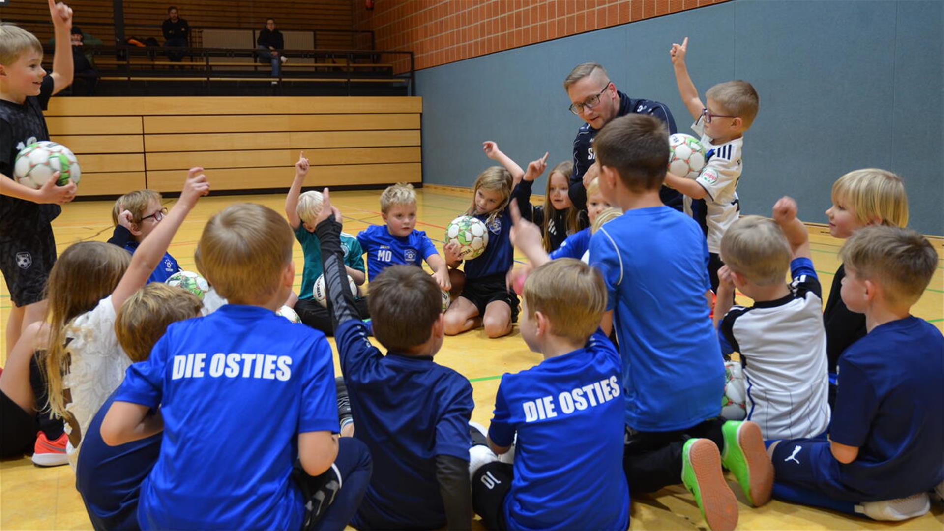 Ein Mann sitzt mit vielen Kindern im Kreis auf dem Boden einer Sporthalle. Ein paar der Kinder haben einen Fußball in der Hand oder melden sich für eine Wortmeldung. 