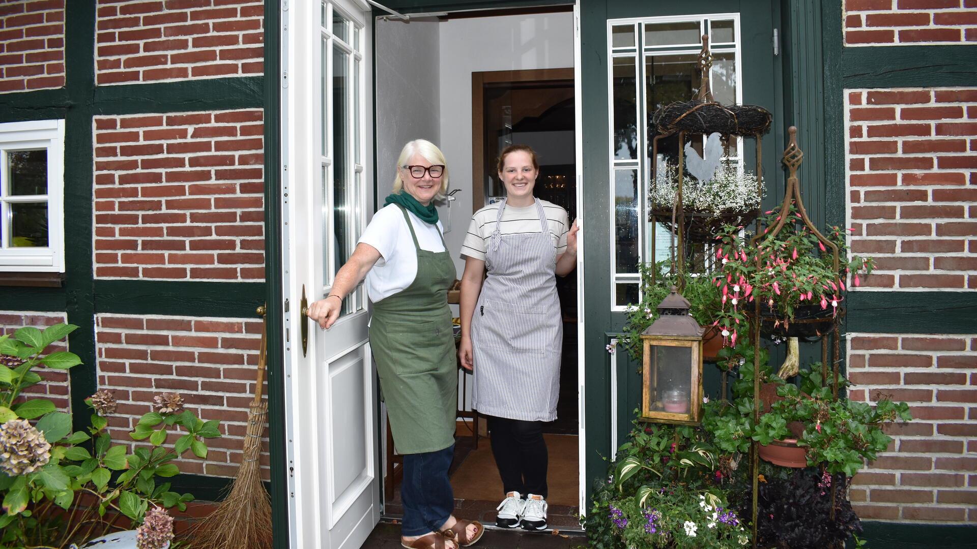 Zwei Frauen in Schürzen stehen in der Eingangtür eines Fachwerkhauses.