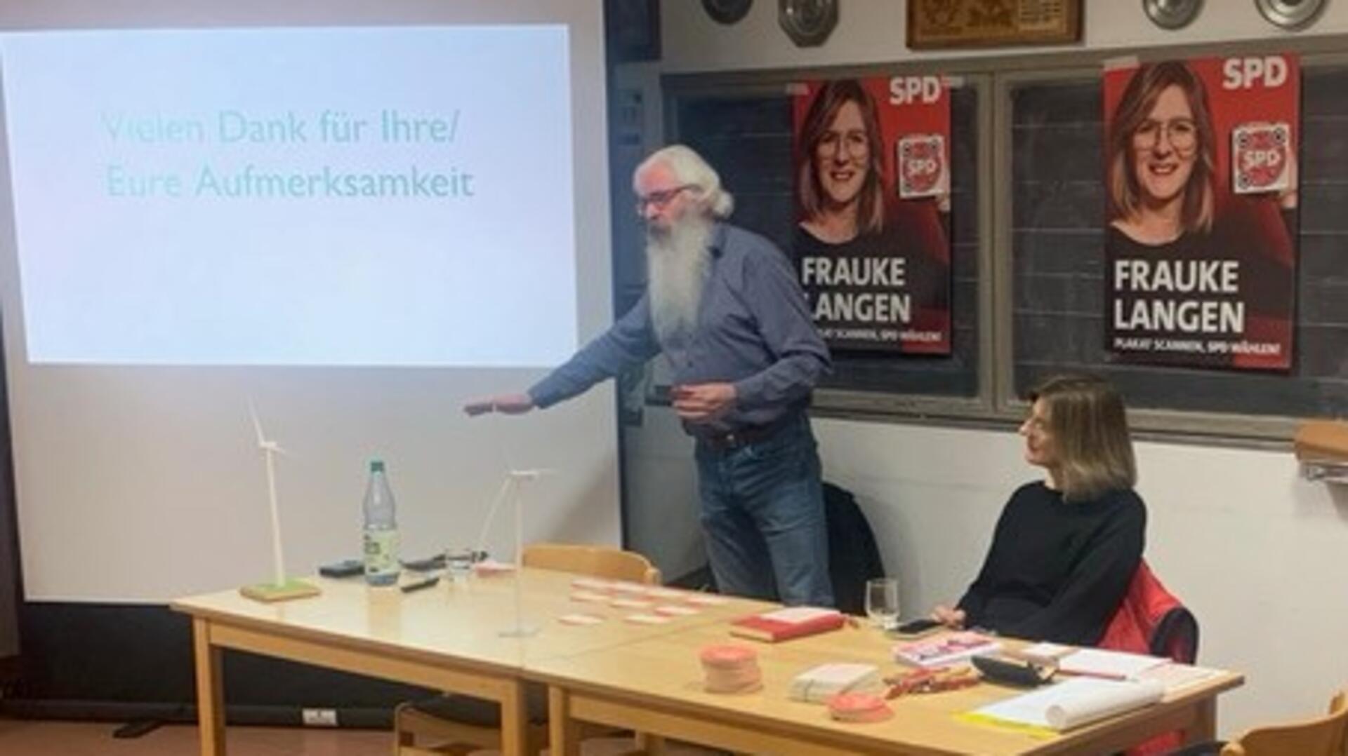 Ein Mann mit weißem Bart steht vor einer Leinwand und hält einen Vortrag über Windenergie. Auf der Leinwand ist der Text „Vielen Dank für Ihre/Eure Aufmerksamkeit“ zu sehen. Neben ihm sitzt eine Frau an einem Tisch, im Hintergrund hängen SPD-Poster mit ihrem Foto und dem Text Frauke Langen.