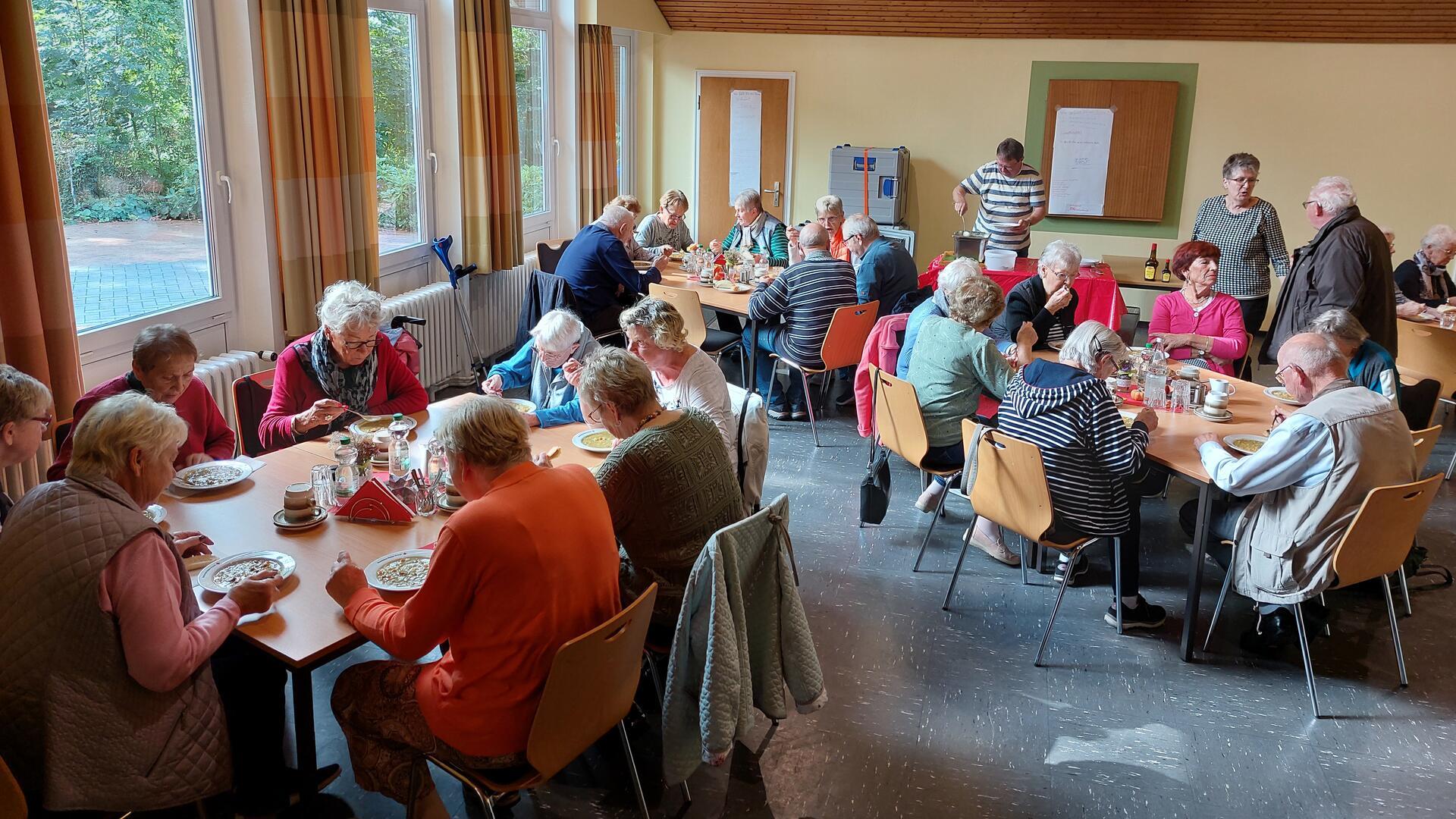 Zahlreiche Menschen sitzen in einem Saal an mehreren Tischen und essen gemeinsam Suppe.