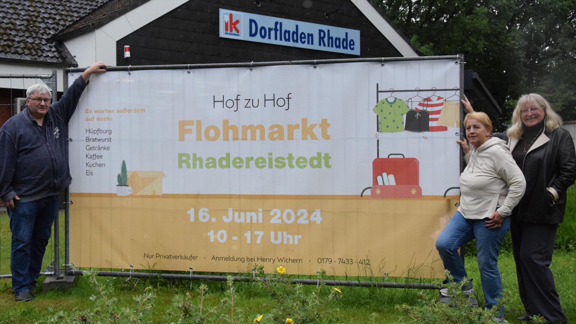 Henry Wichern vom Organisationsteam sowie Gisela Marquarding und Birgit Arndt (rechts) von den Rhader Landfrauen am Banner, das im Rhader Ortskern am Dorfladen für den Flohmarkt in Rhadereistedt wirbt.