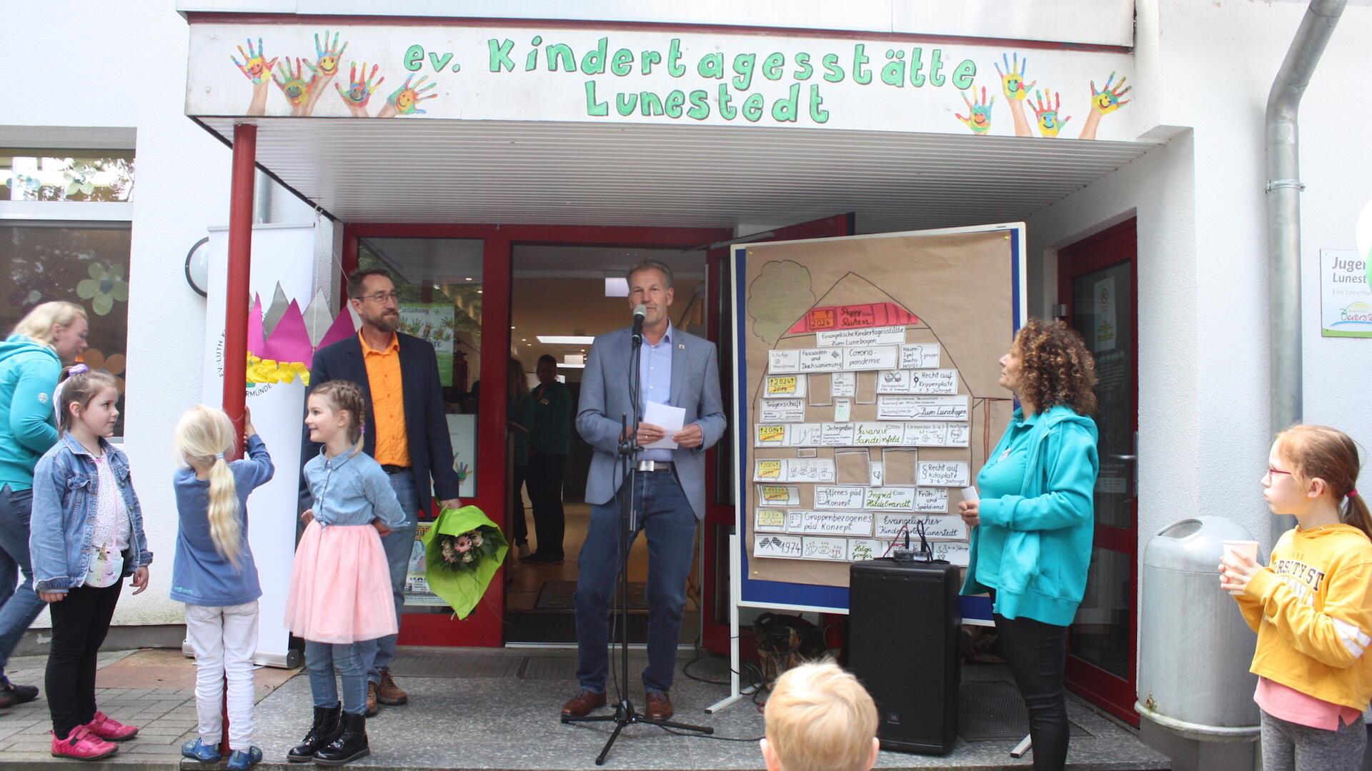 Einige Kinder und Erwachsene stehen vor einem Eingang, über dem "ev. Kindertagesstätte Lunestedt" steht. Ein Mann steht an einem Mikrofon und hält eine Rede.